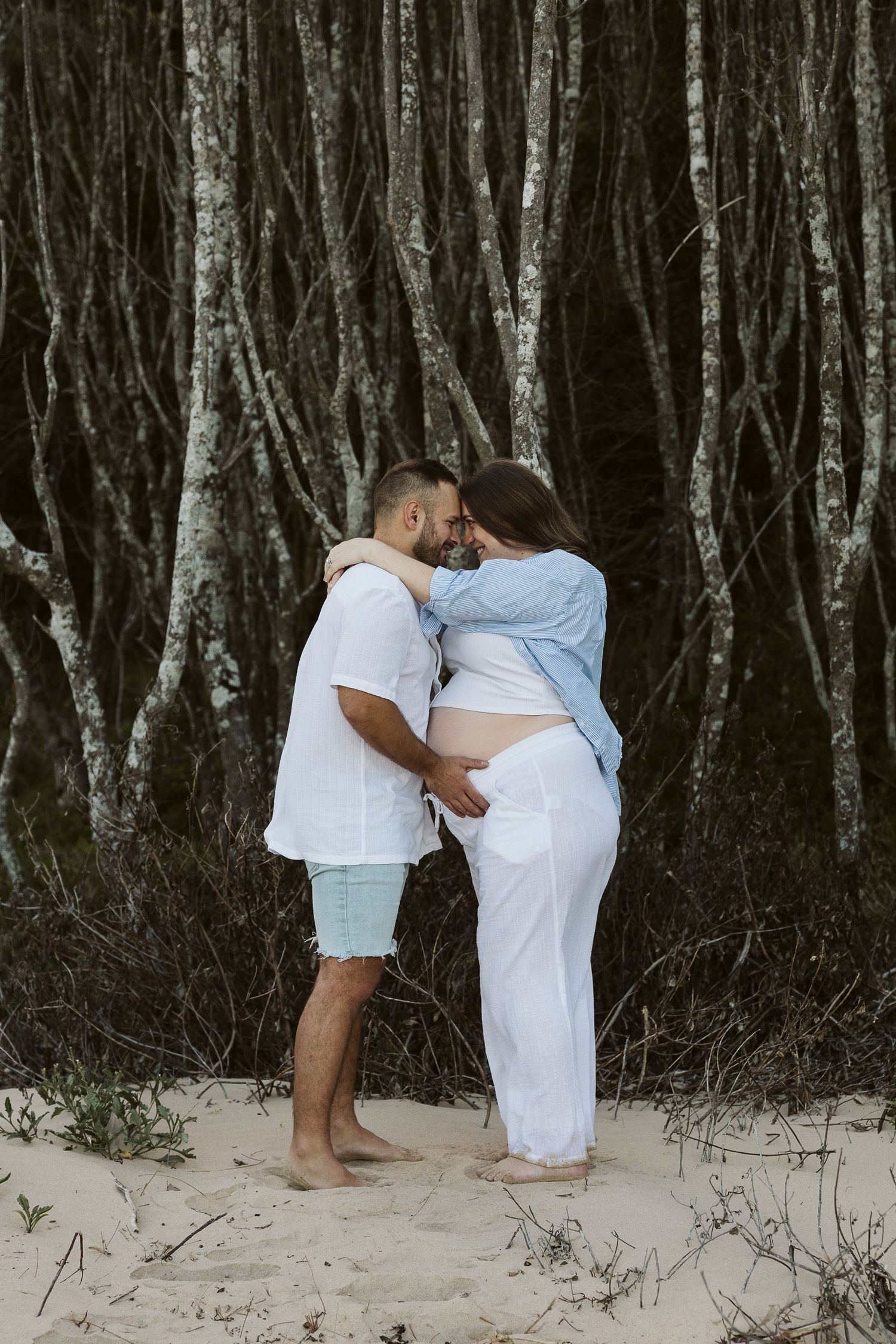 Couple sharing intimate forehead touch near dark trees on sandy beach, pregnant woman in white pants.