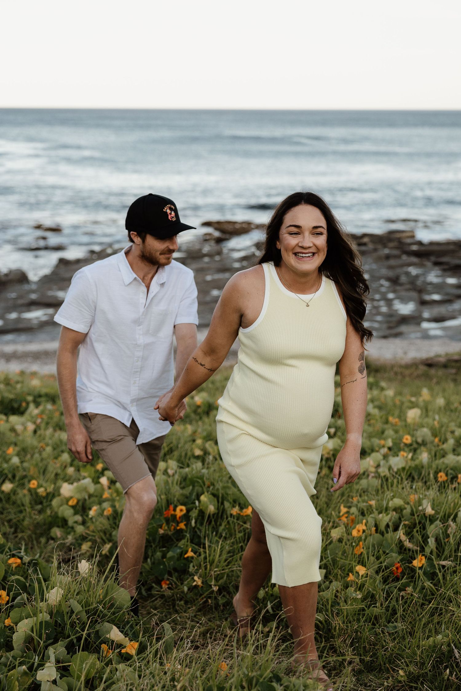 Happy pregnant woman in yellow dress holds partners hand near ocean wildflowers.