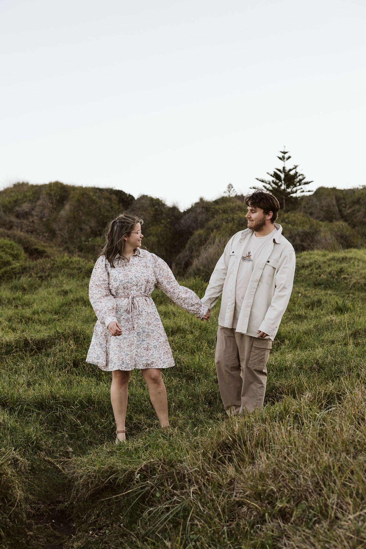 Couple holding hands and walking through lush green hillside, smiling at each other at dusk.