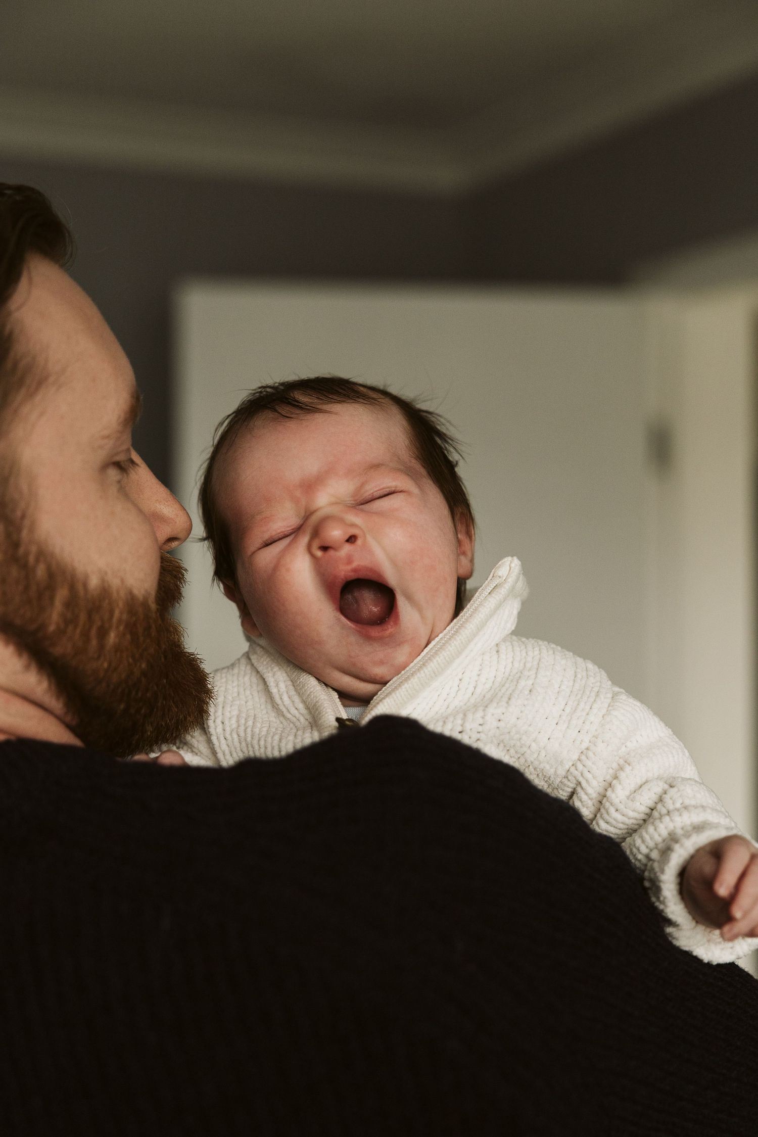 Bearded father holding yawning newborn baby dressed in white knit outfit.