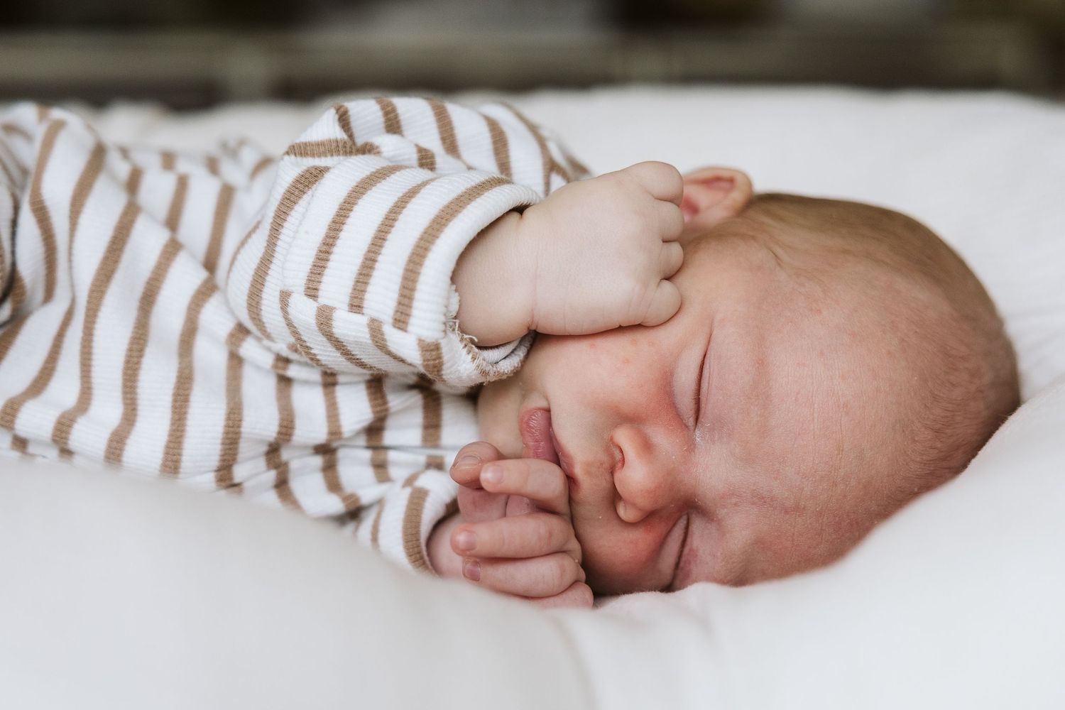 Sleeping newborn baby in brown striped onesie with hands tucked under cheek on white surface.