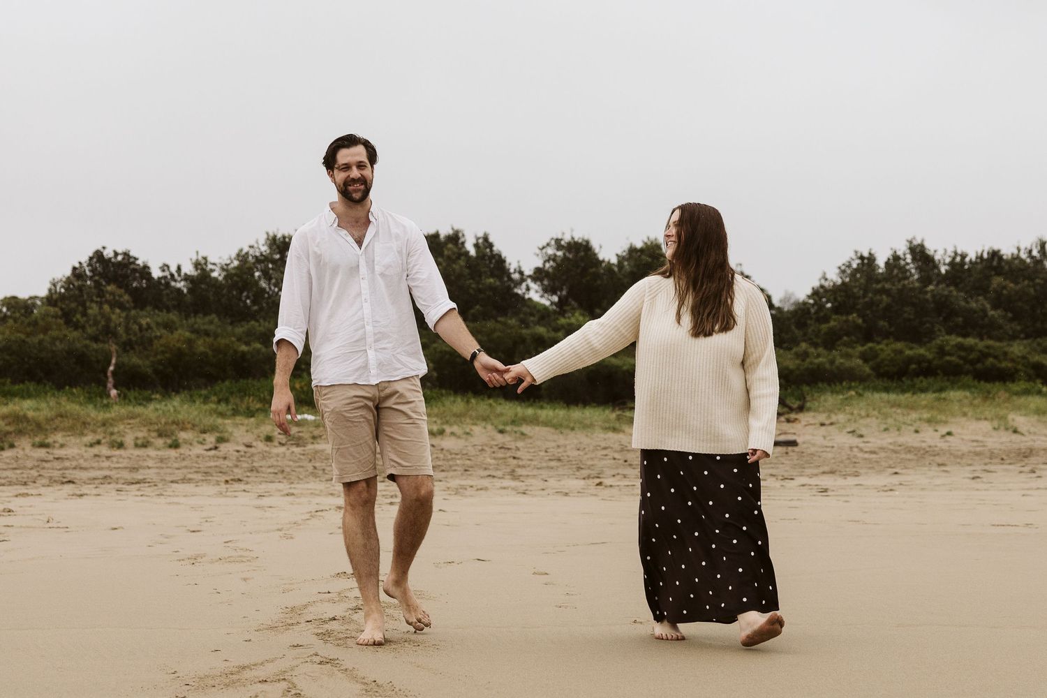 Happy couple holding hands and walking barefoot on sandy beach together.