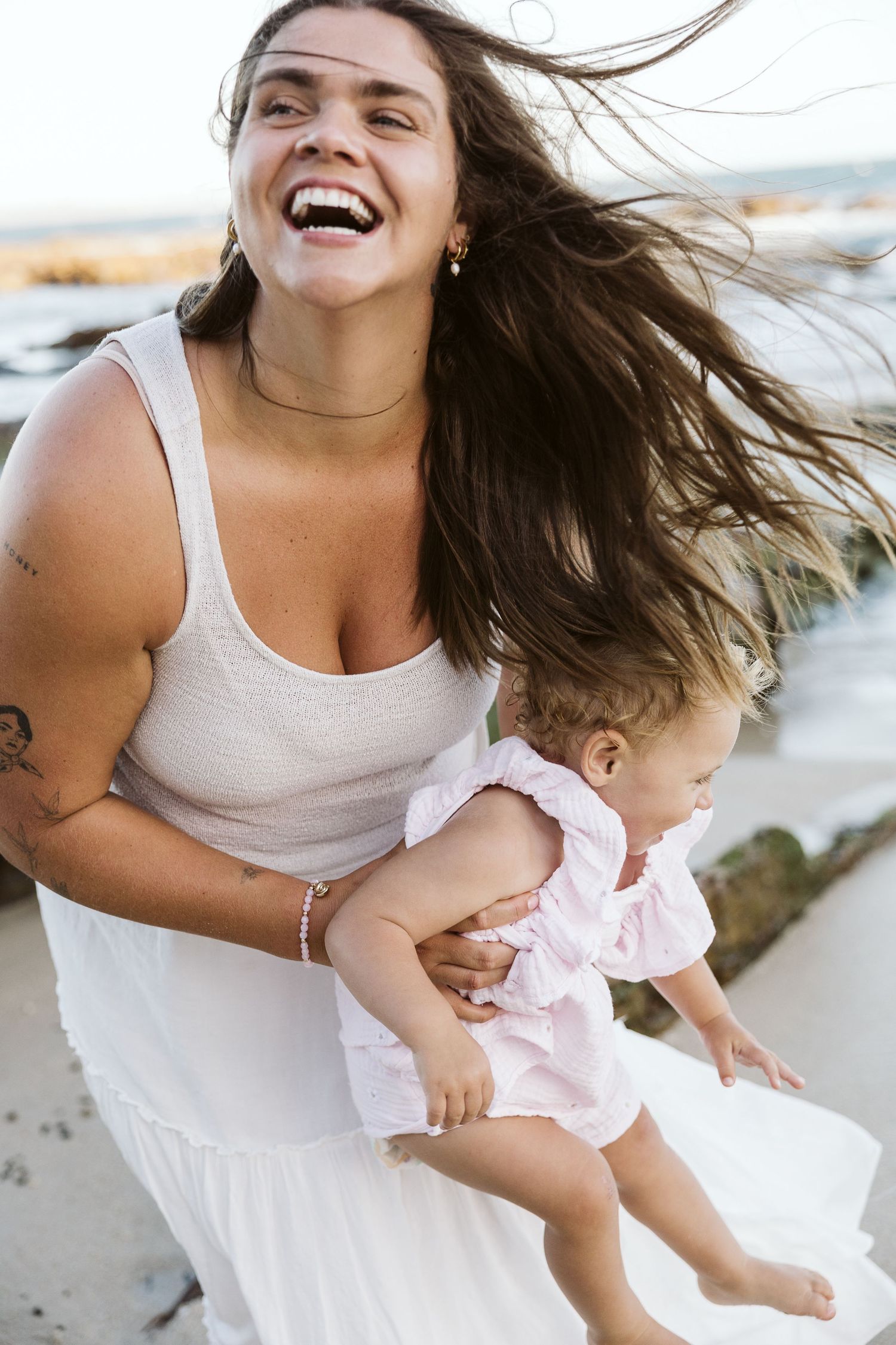 A laughing woman in a white dress holds a baby girl at the beach, her hair blowing in the wind.