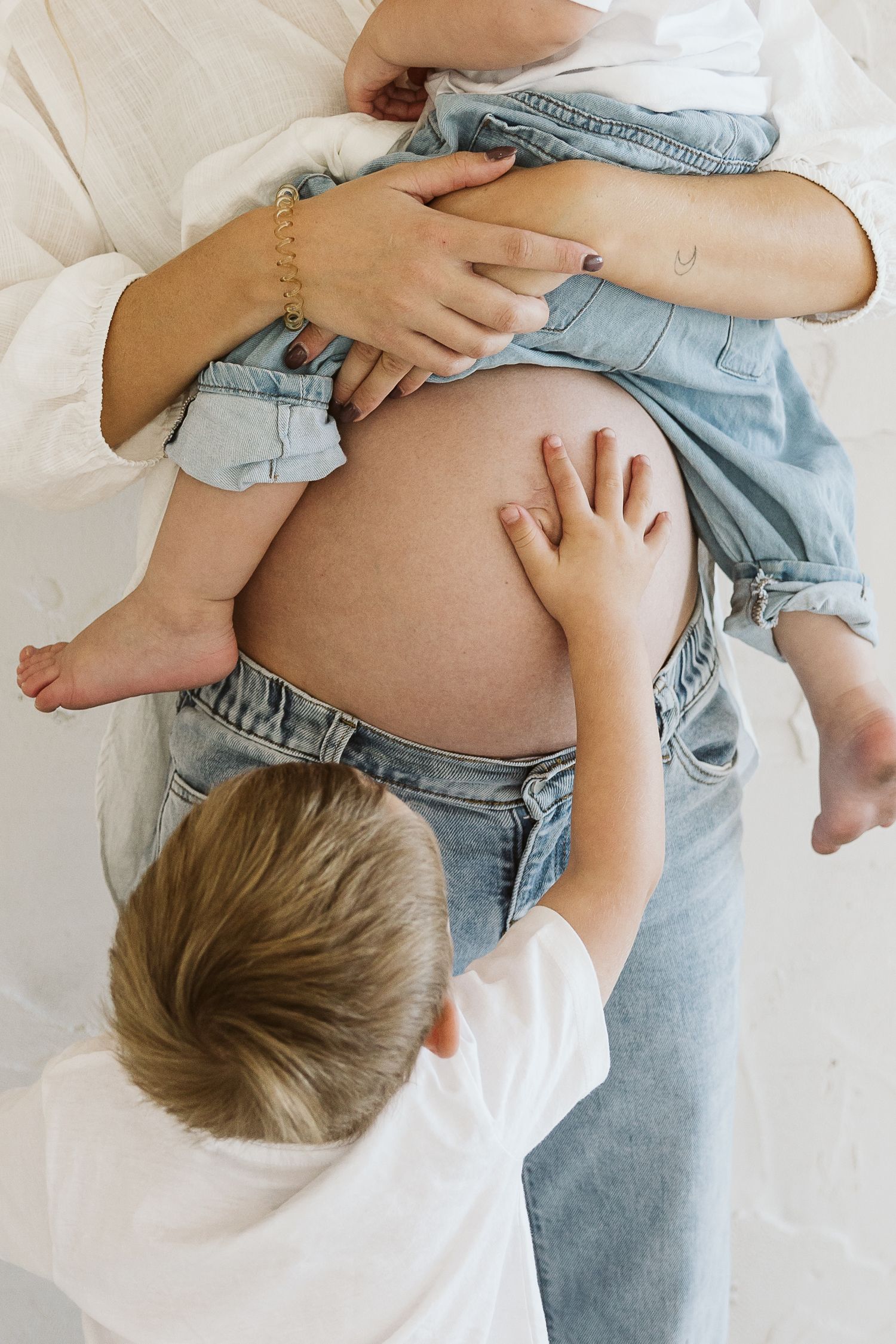 Child touches pregnant mothers bare belly while she holds a baby dressed in denim against white background.