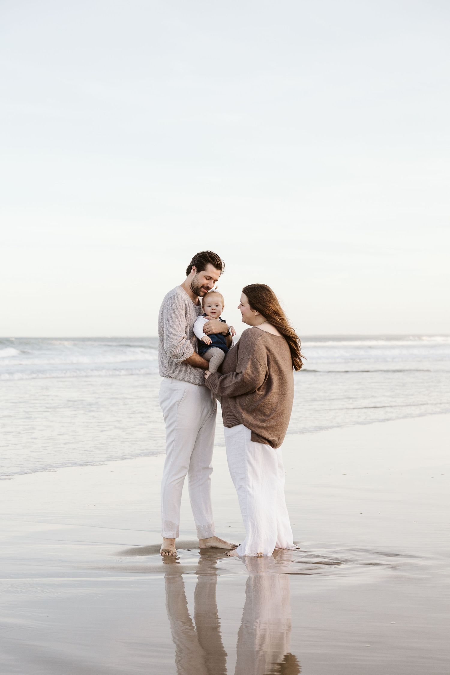 A young family with a baby stands barefoot on a reflective wet beach, sharing a tender moment by the ocean waves.