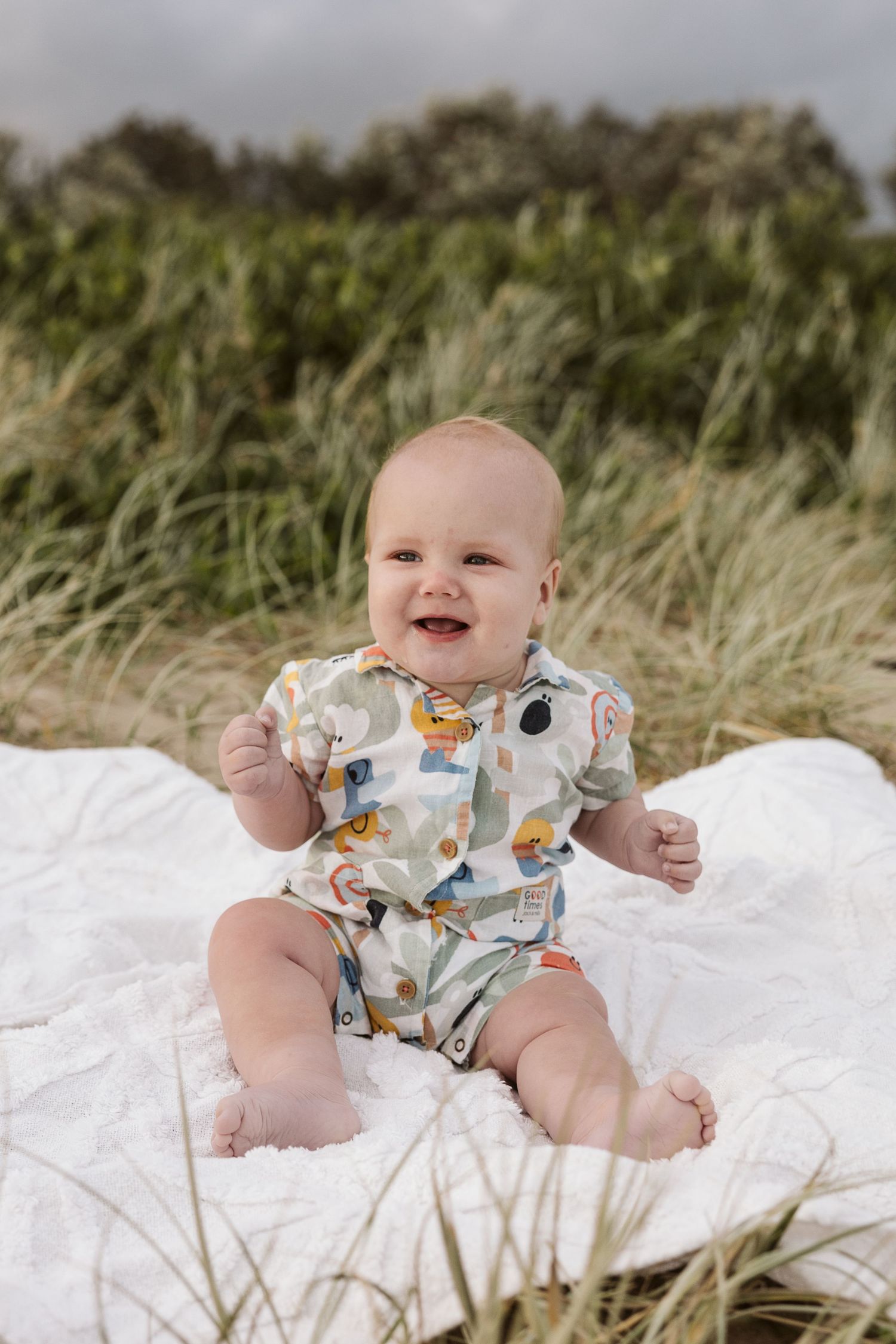 Smiling baby in a colorful printed romper sitting on a white blanket outdoors among green grass.