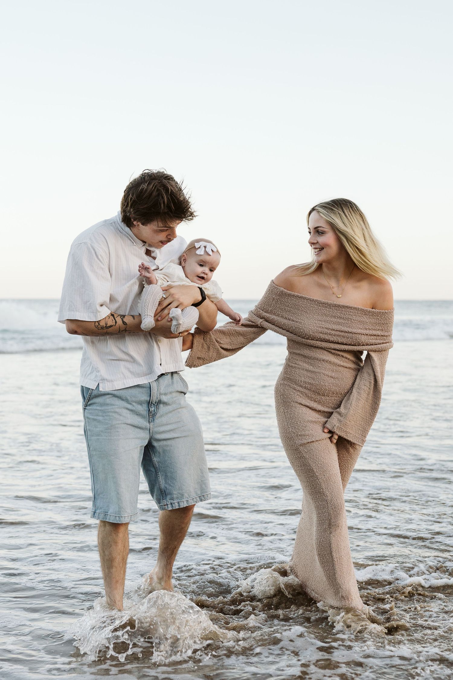 Young family wading in ocean waves at sunset, father holding baby while mother smiles nearby.