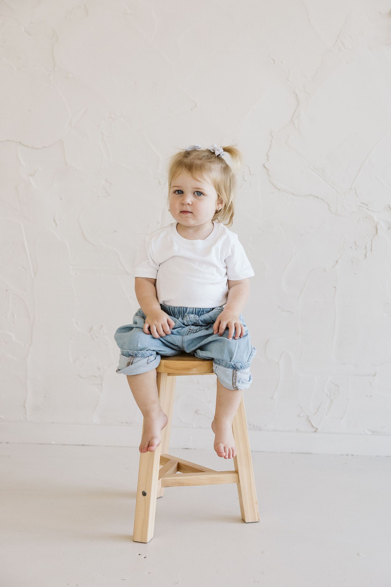 Cute toddler girl with blonde pigtail sits cross-legged on wooden stool wearing white tee and rolled denim jeans.