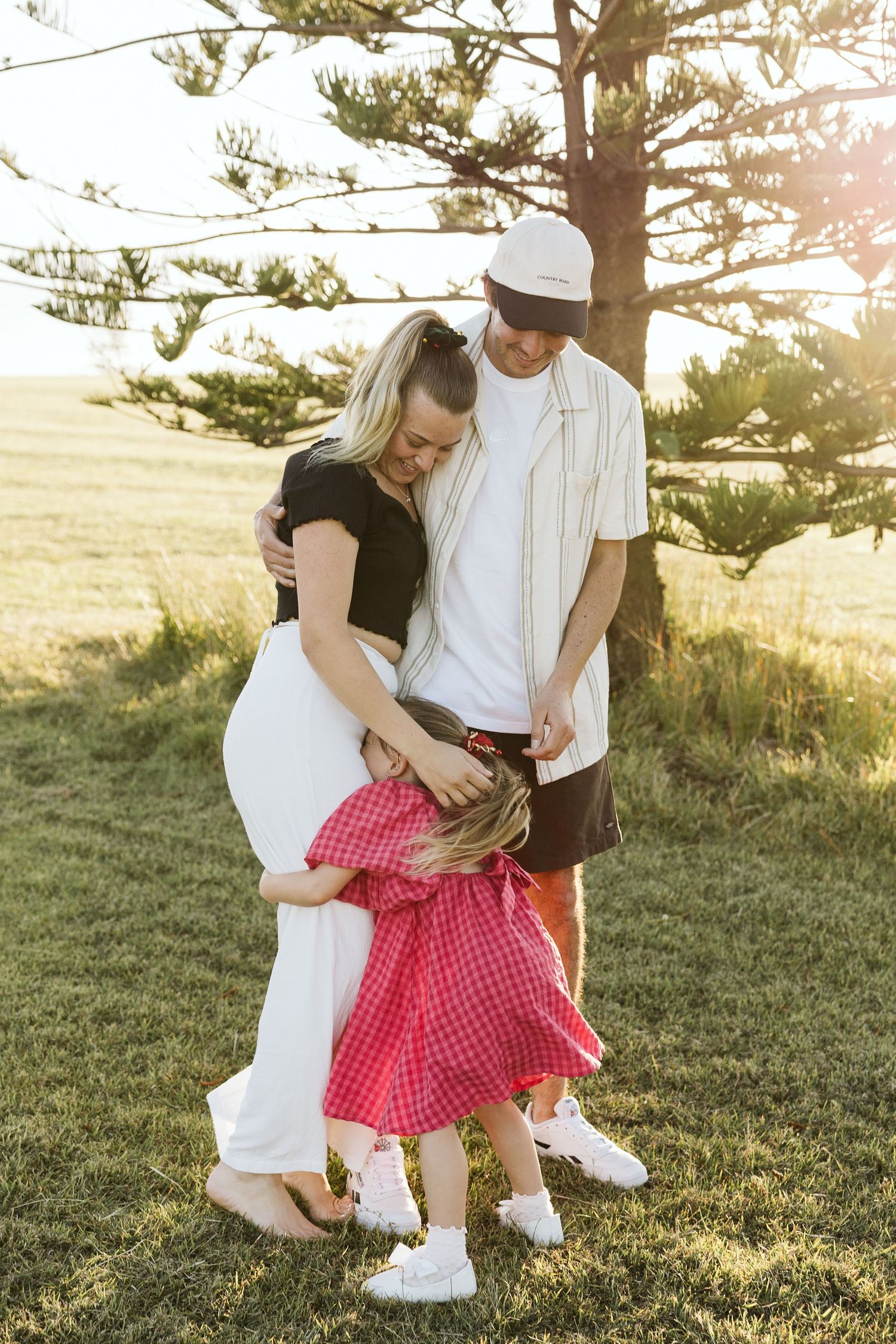 A happy family of three sharing a warm embrace outdoors on green grass with a pine tree in golden sunlight.