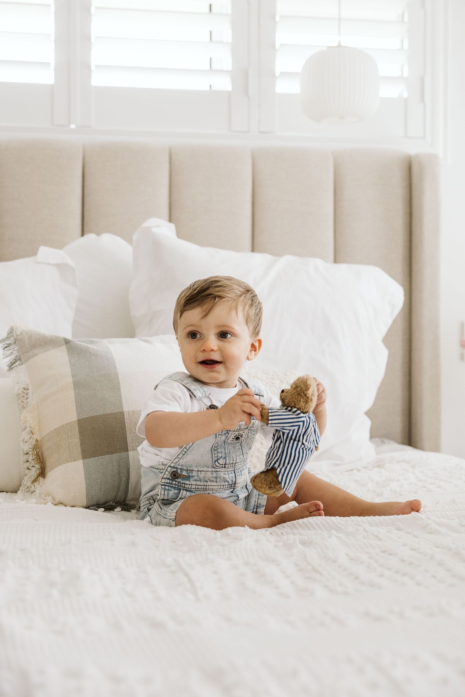 Smiling toddler boy in denim overalls holding a teddy bear, sitting on a white bed with pillows.
