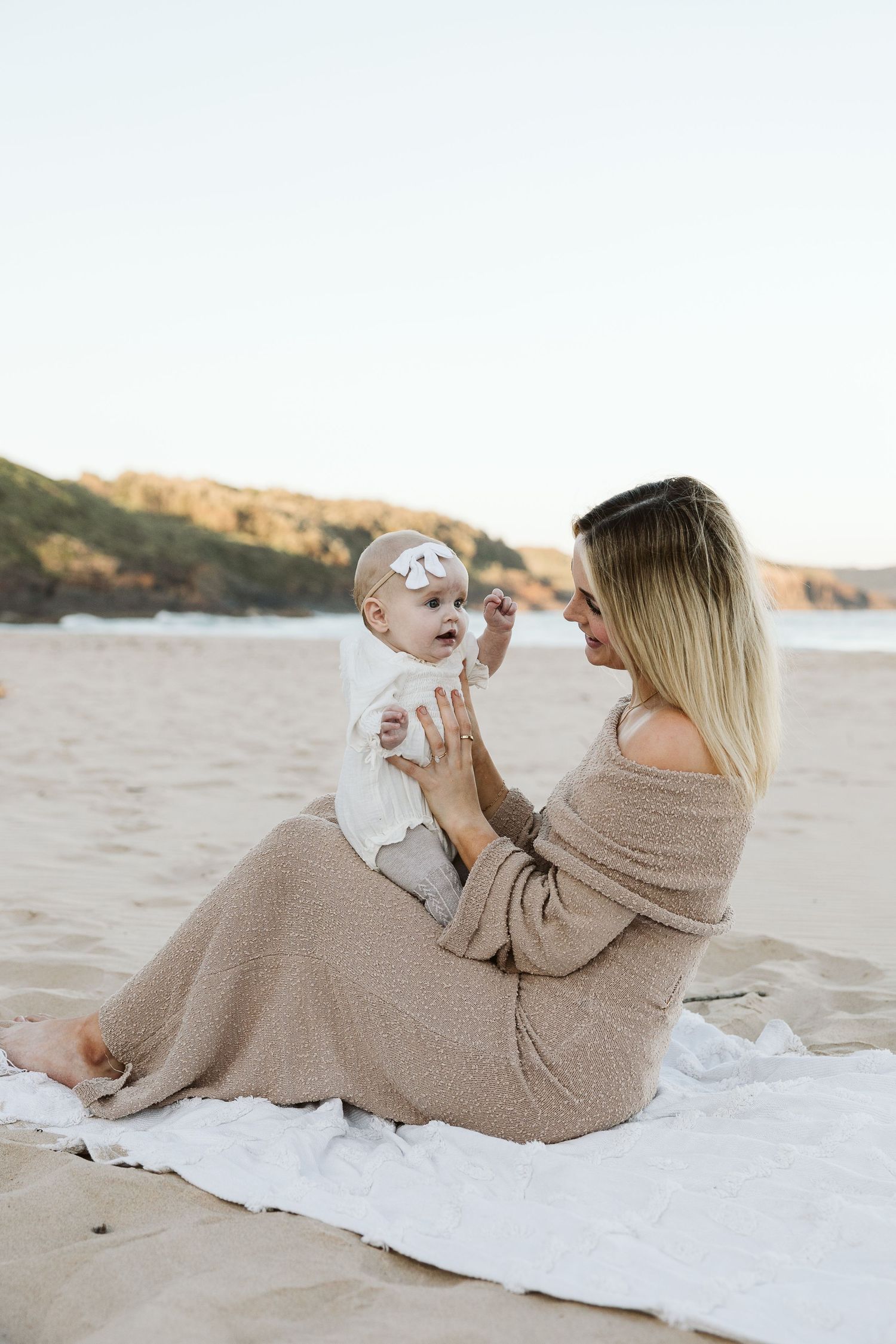 A mother sits on a white blanket at the beach, lovingly holding her baby girl in a white outfit with a bow headband.
