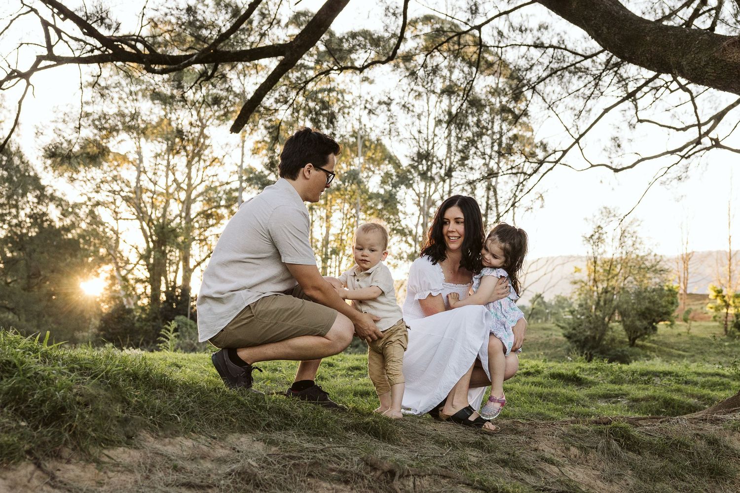 Family of four enjoying a golden sunset in a lush park, parents holding toddlers near a tree.