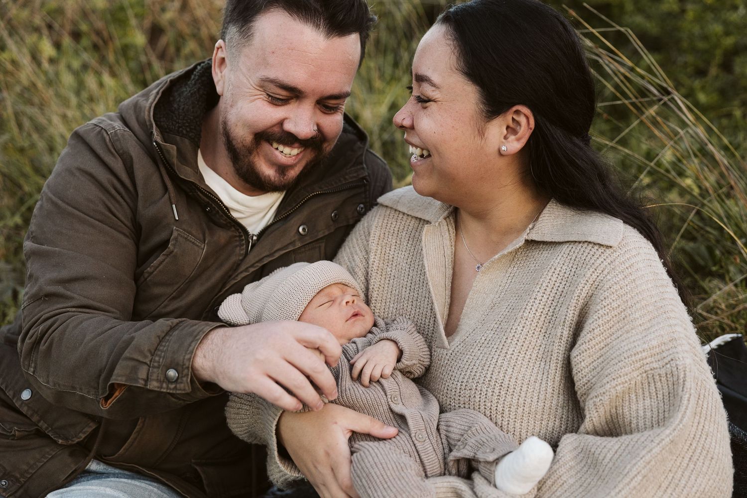 Smiling couple sitting outdoors in tall grass, lovingly looking at their newborn baby.