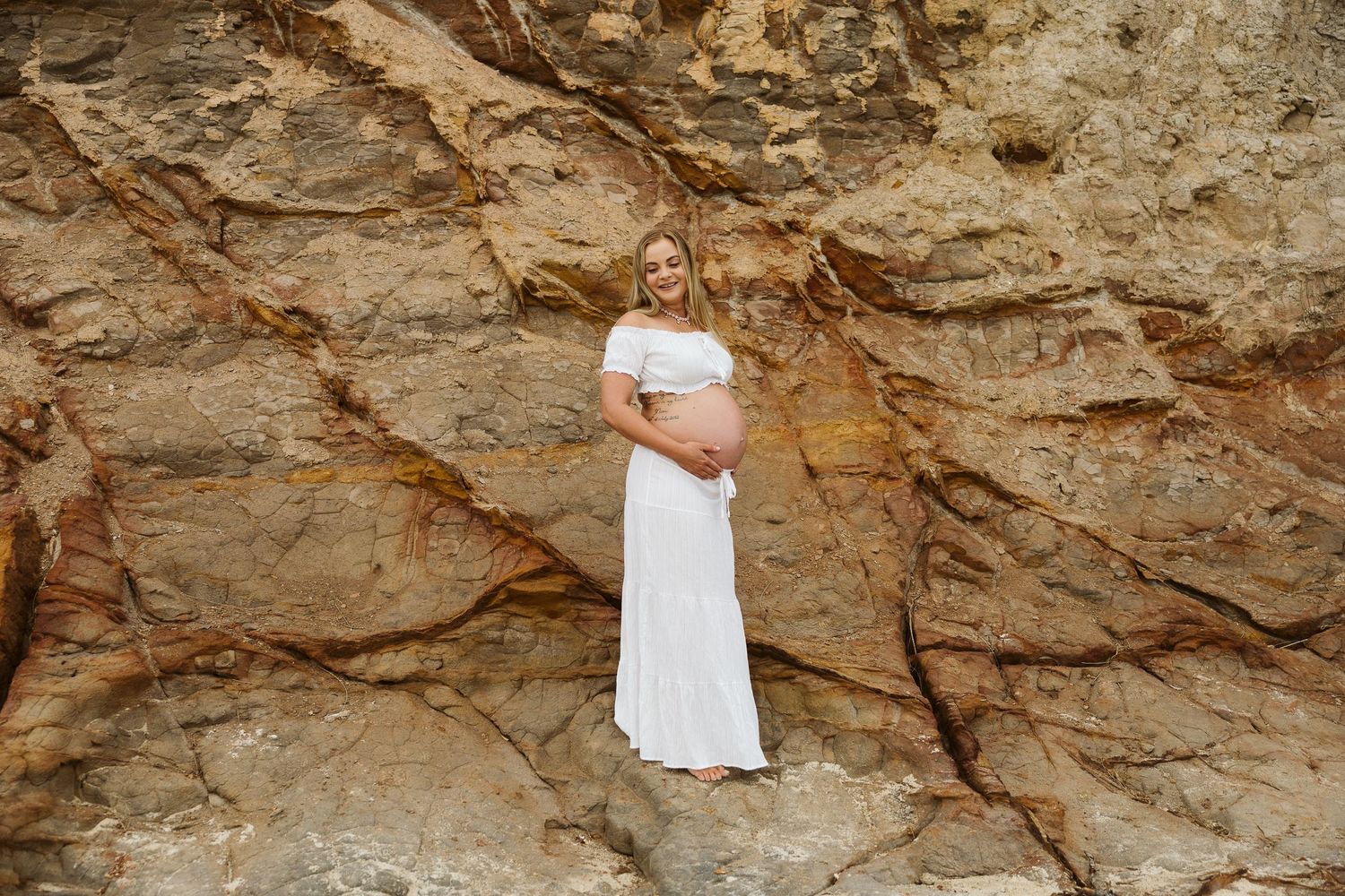Pregnant woman in white off-shoulder dress posing against textured golden rock face outdoors.