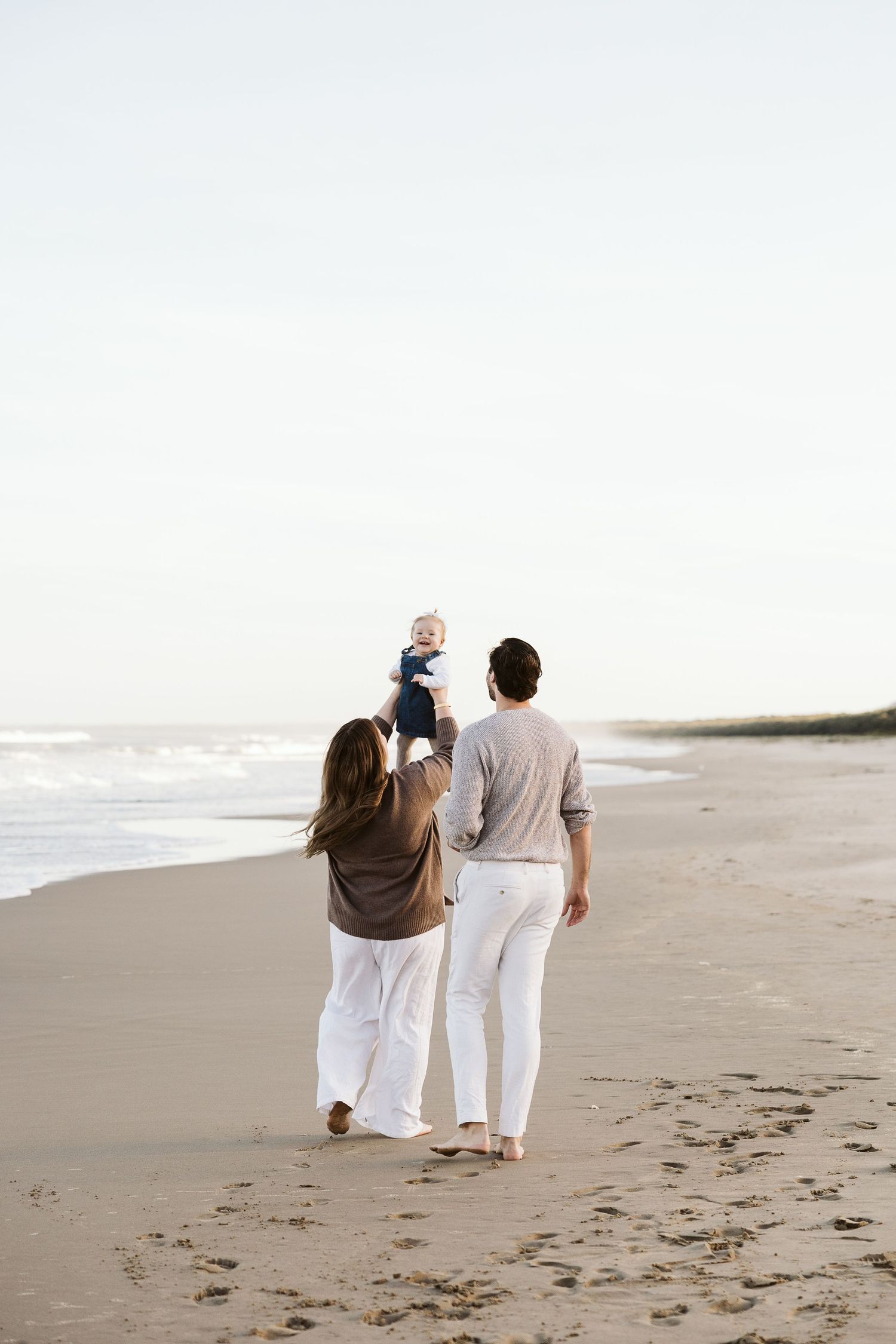 Parents walking barefoot on beach holding baby up in the air near ocean waves.