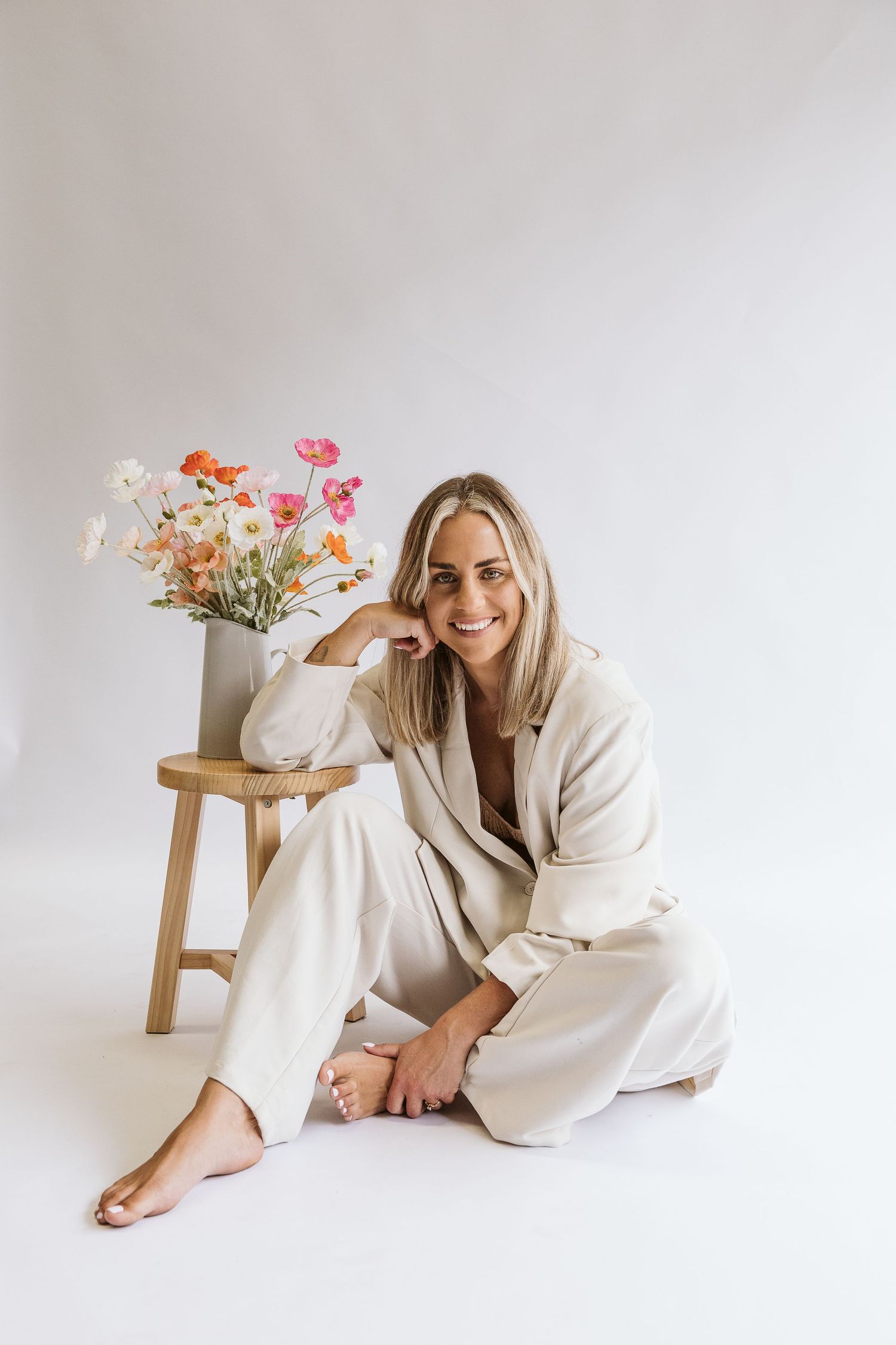 Smiling blonde woman in white linen suit sits on floor beside colorful flowers.