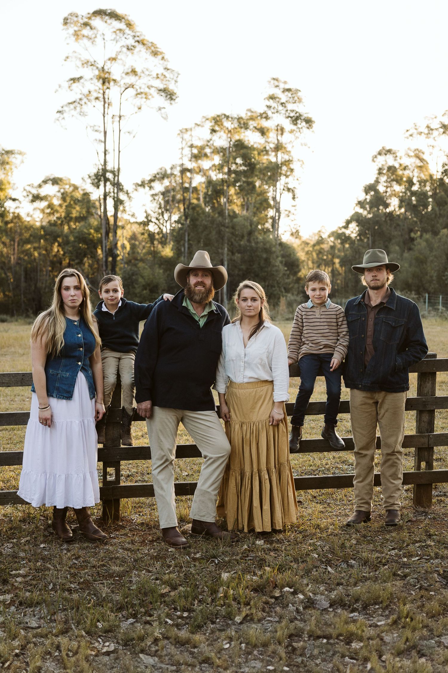 Family of six posing by a rustic wooden fence in a golden field at sunset wearing country-style outfits.
