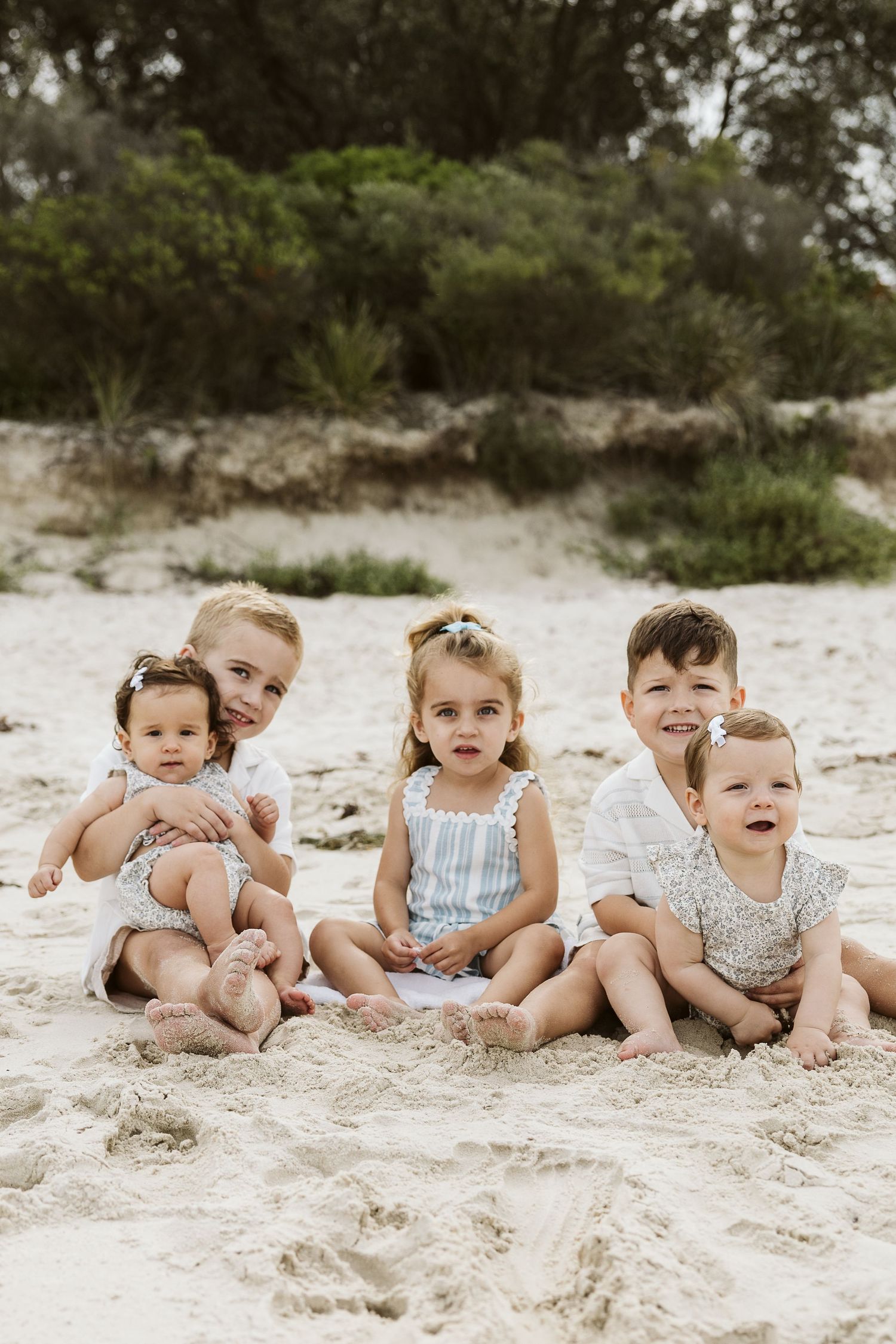 Five young children sitting together on sandy beach with lush green trees in background.