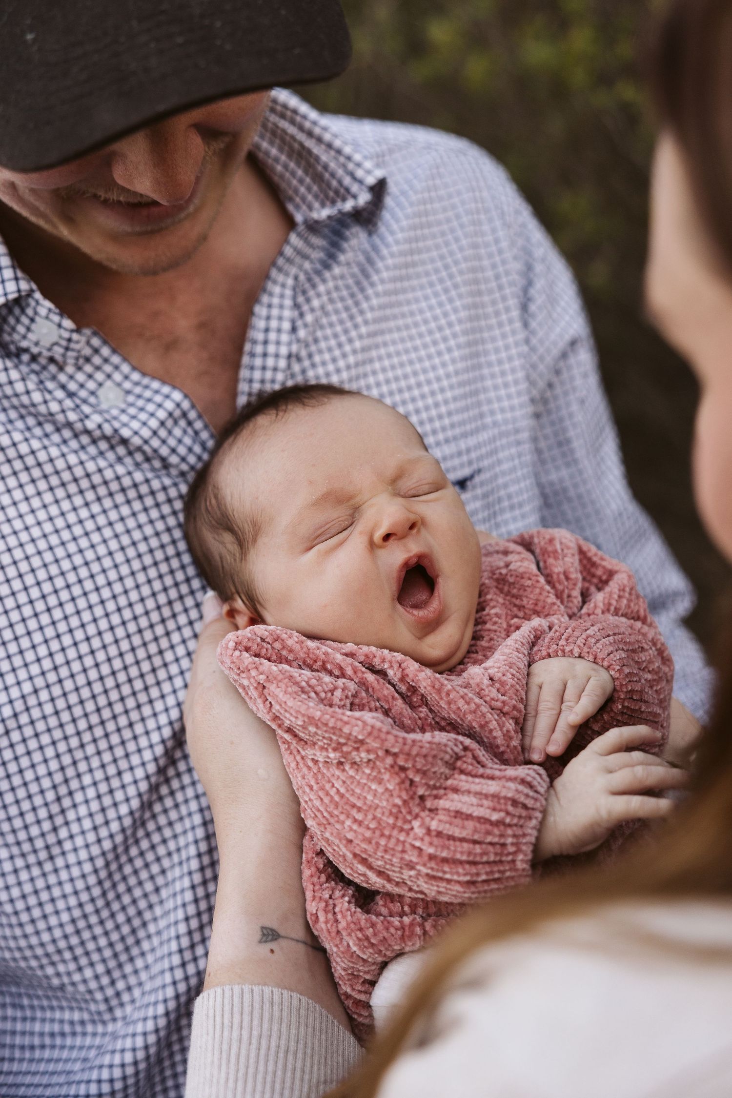 Newborn baby yawning while being held by father in plaid shirt and cap, mother watching lovingly.