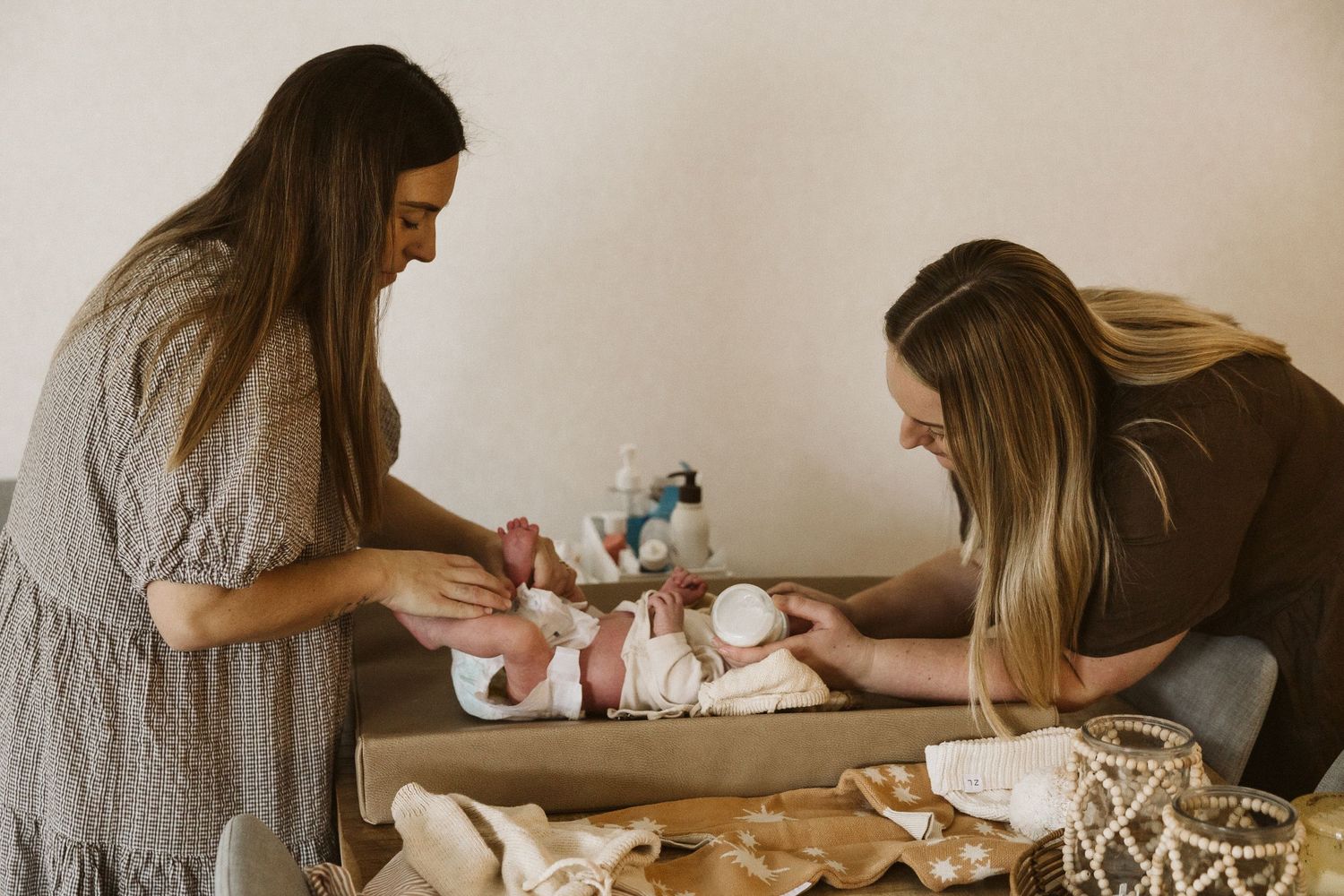 Two mothers changing newborn baby on changing table with nursery supplies nearby.