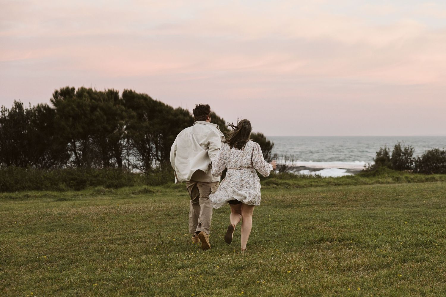 Couple running hand-in-hand across green coastal field at sunset with ocean and trees behind.