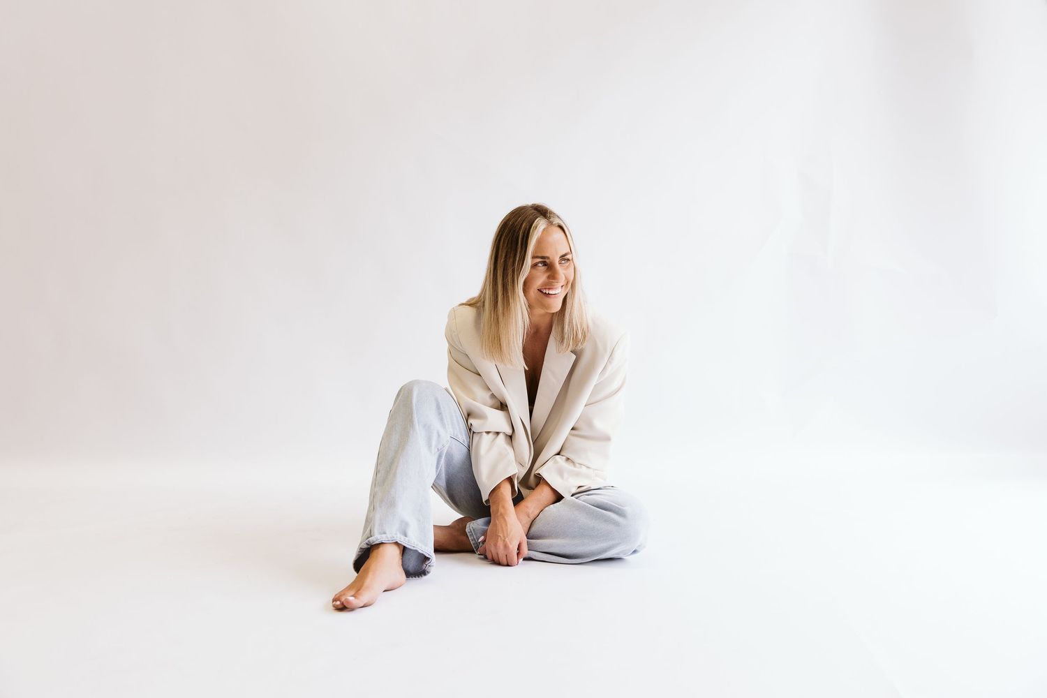 Relaxed blonde woman in beige blazer and jeans sits cross-legged on white floor.