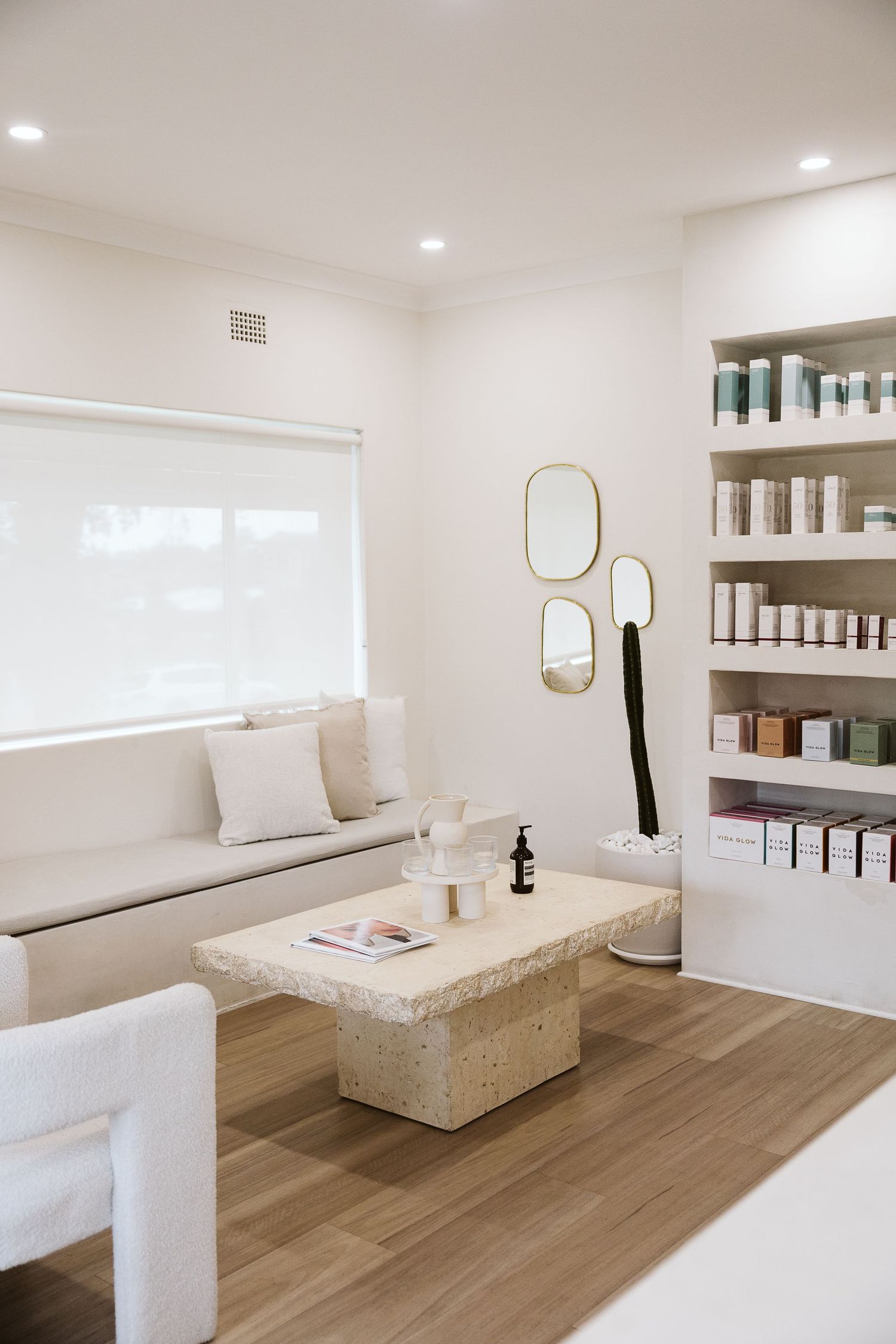 Bright minimalist aesthetics clinic waiting room with travertine coffee table, product shelves, and gold accent mirrors.