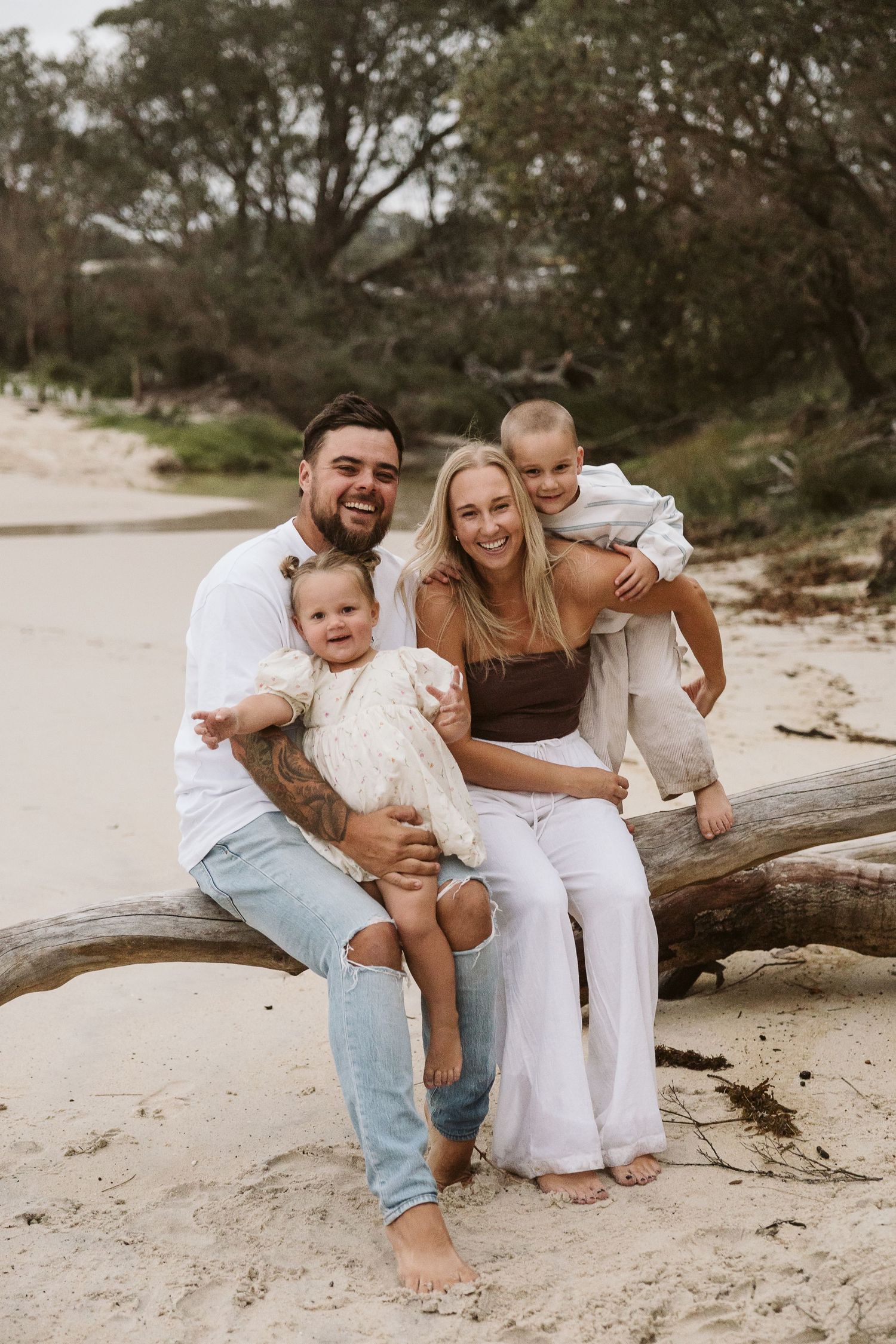 Happy family of four sitting on driftwood log on sandy beach surrounded by trees.