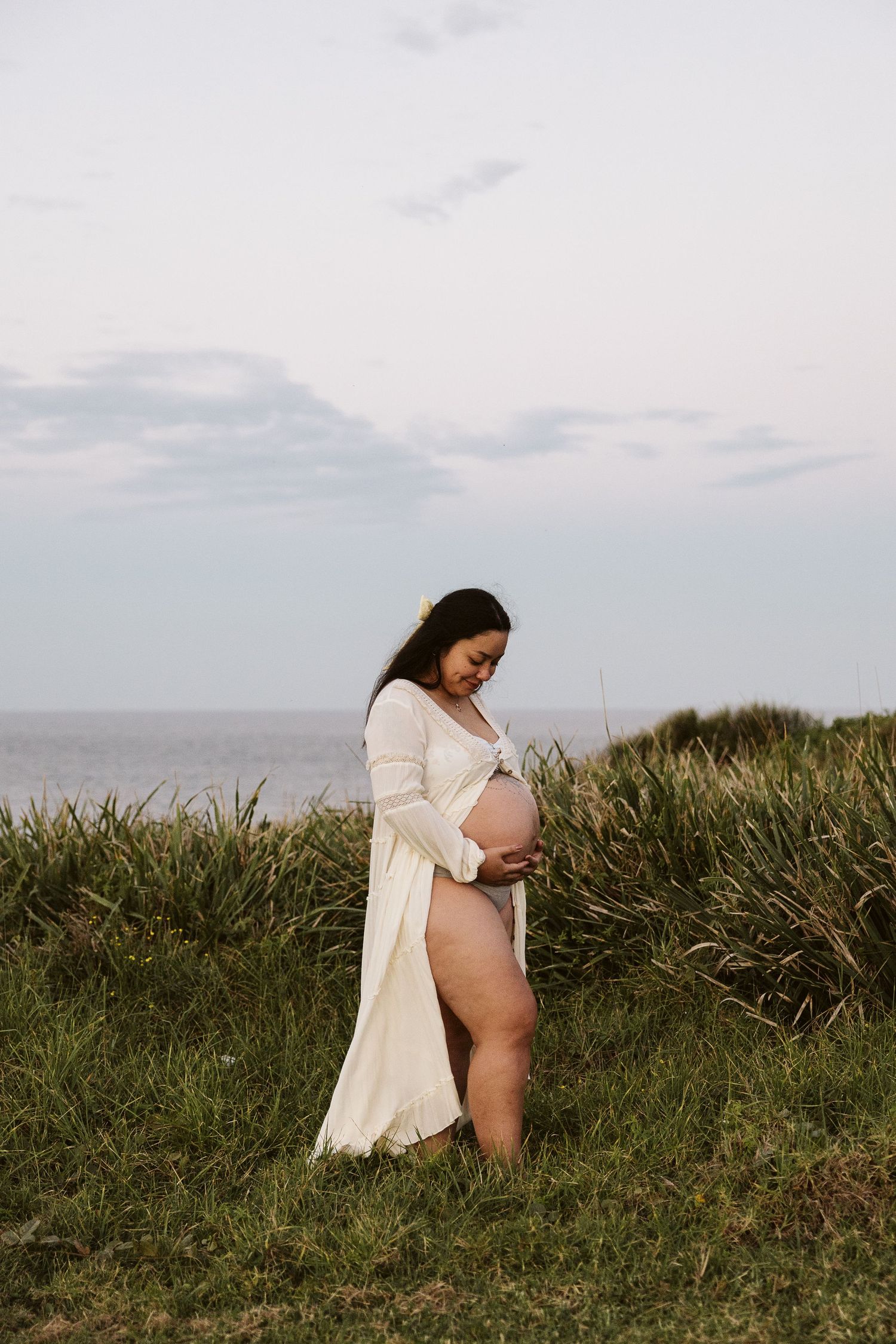 Pregnant woman in white open robe cradling bare baby bump while standing in coastal grass near the ocean.