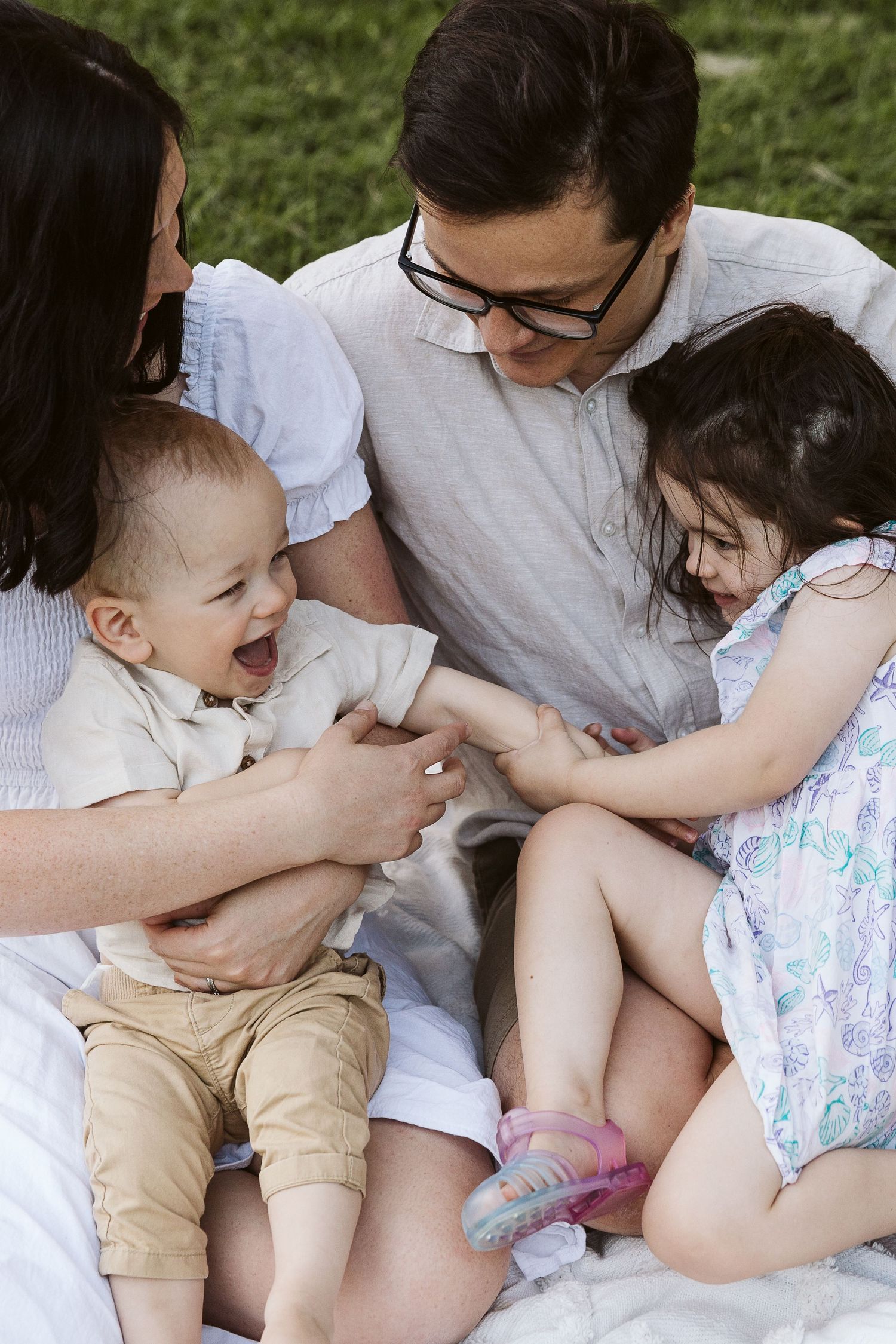 Happy family of four enjoying a sunny outdoor picnic on a white blanket, with parents playing with laughing baby and young girl.