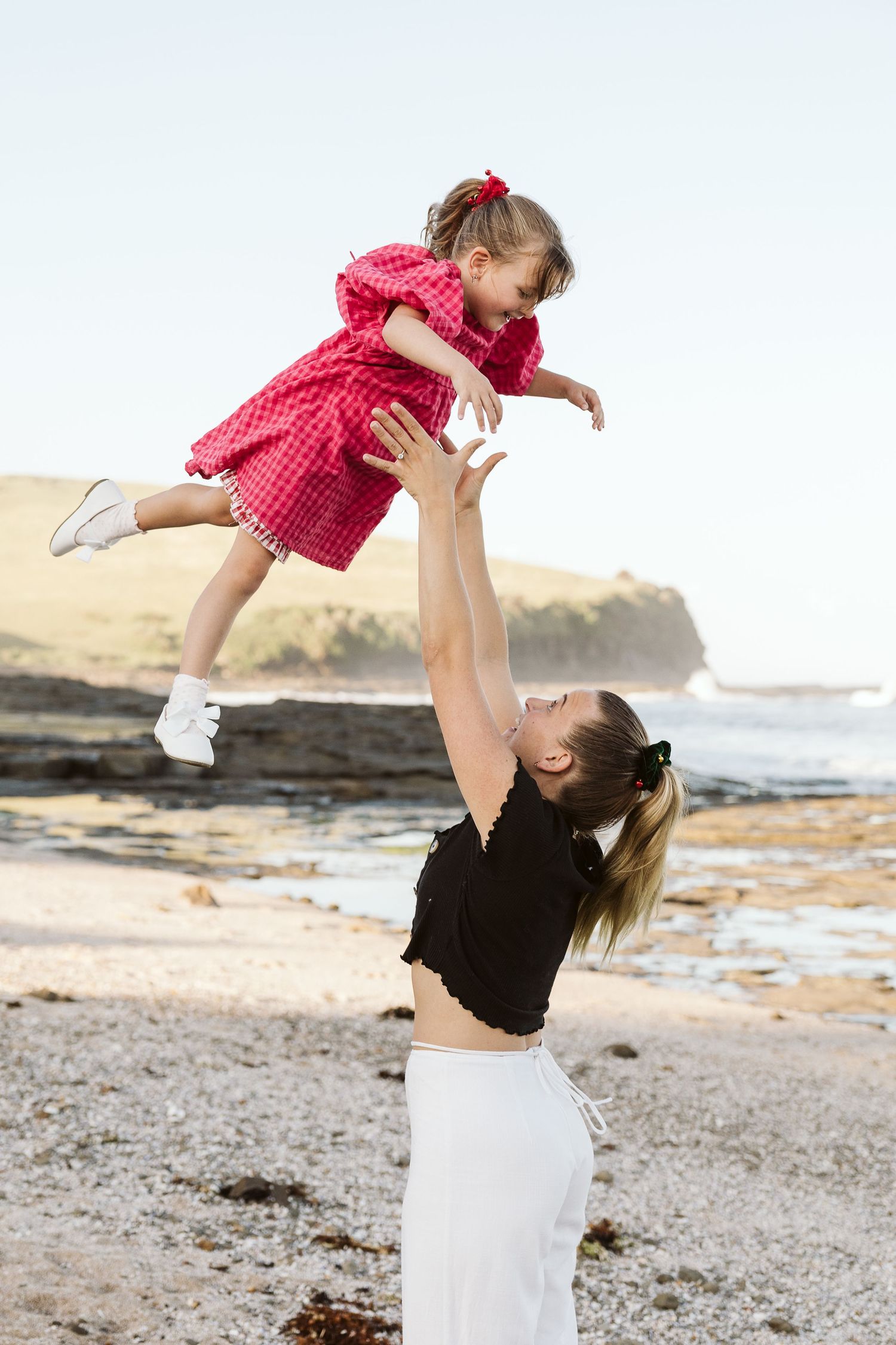 Mother joyfully lifting her young daughter in a pink dress high into the air on a scenic rocky beach.
