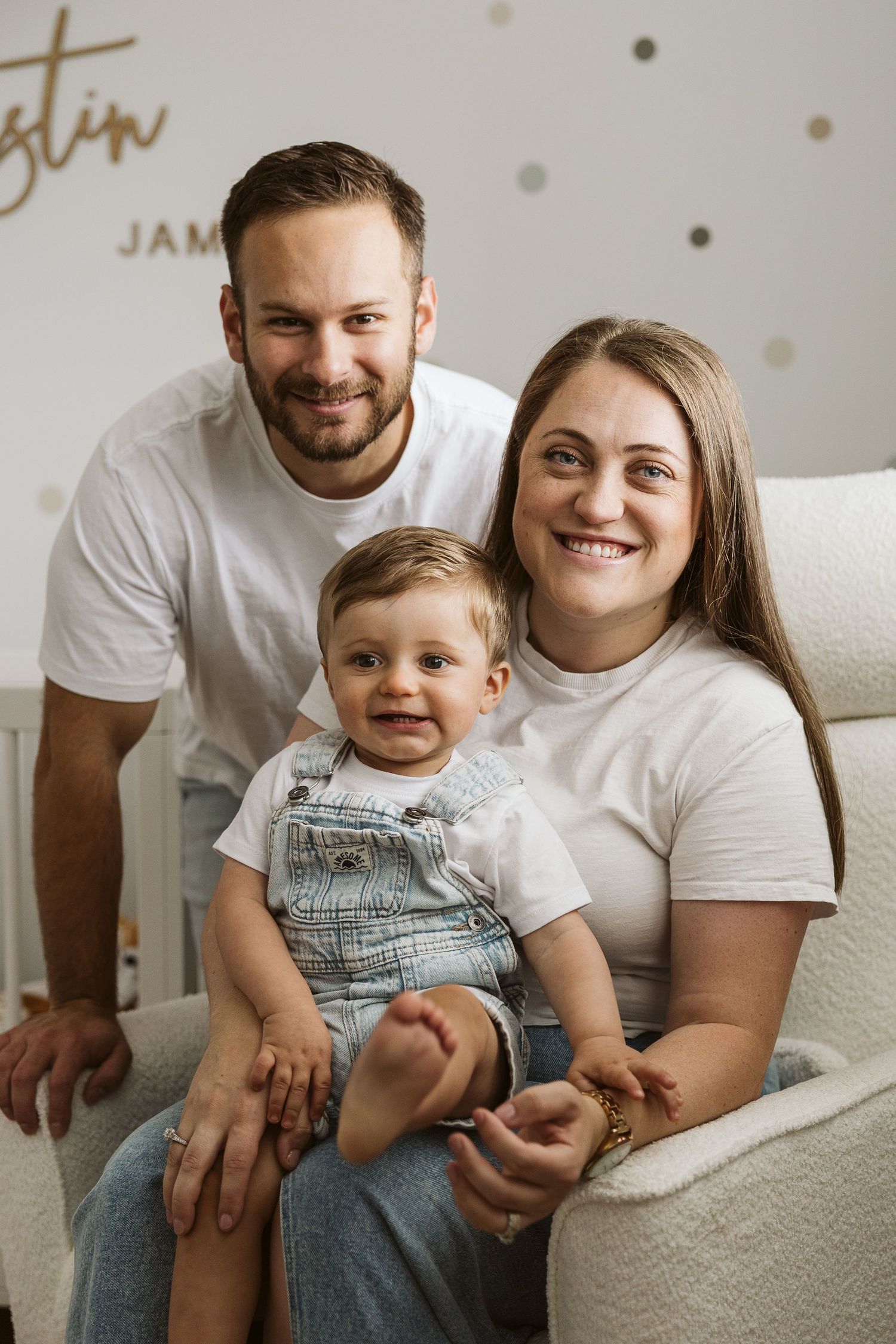 Happy family portrait with parents in white tees and baby in denim overalls sitting in nursery chair.