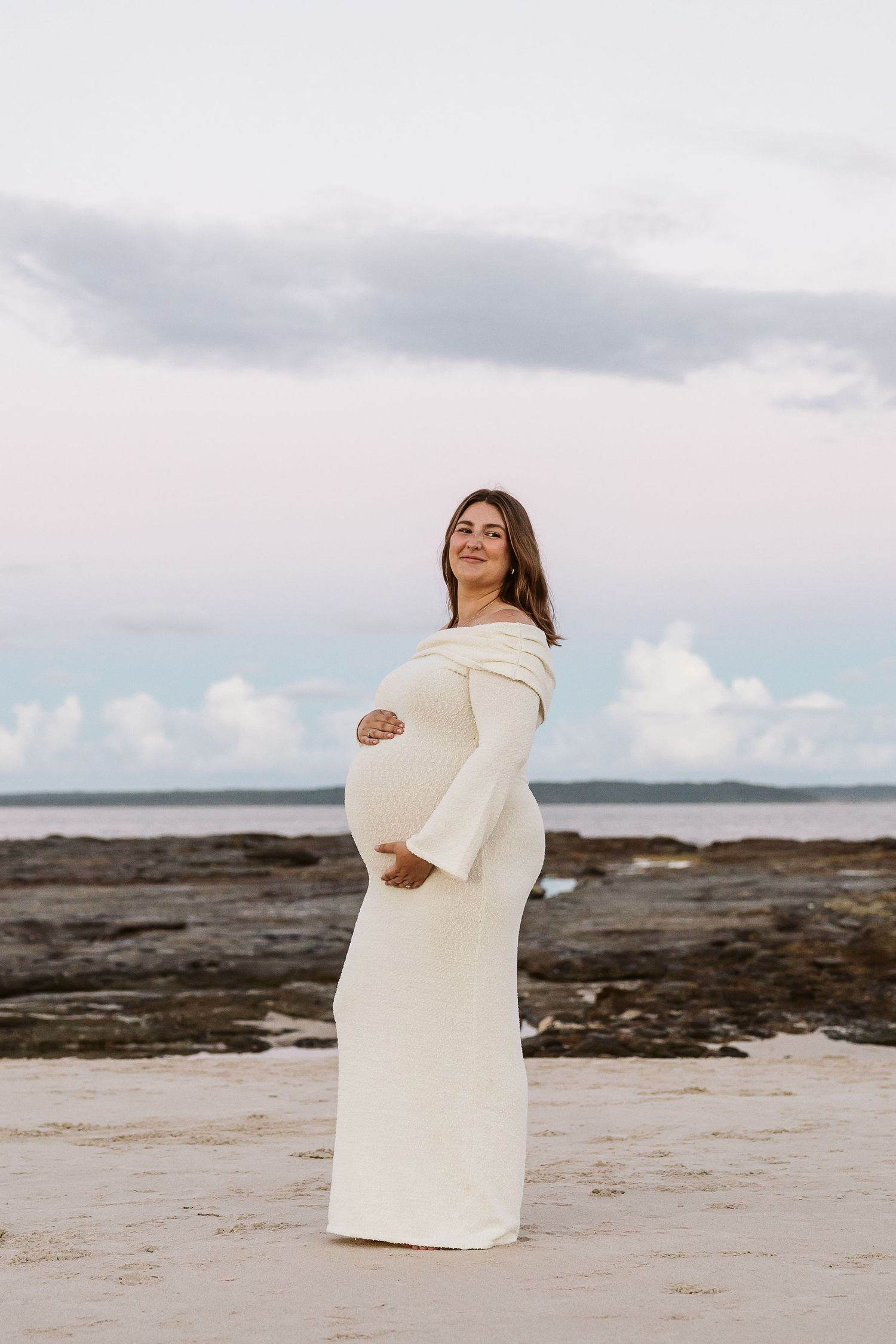 Pregnant woman in cream off-shoulder knit dress stands sideways on beach cradling bump under cloudy sky.