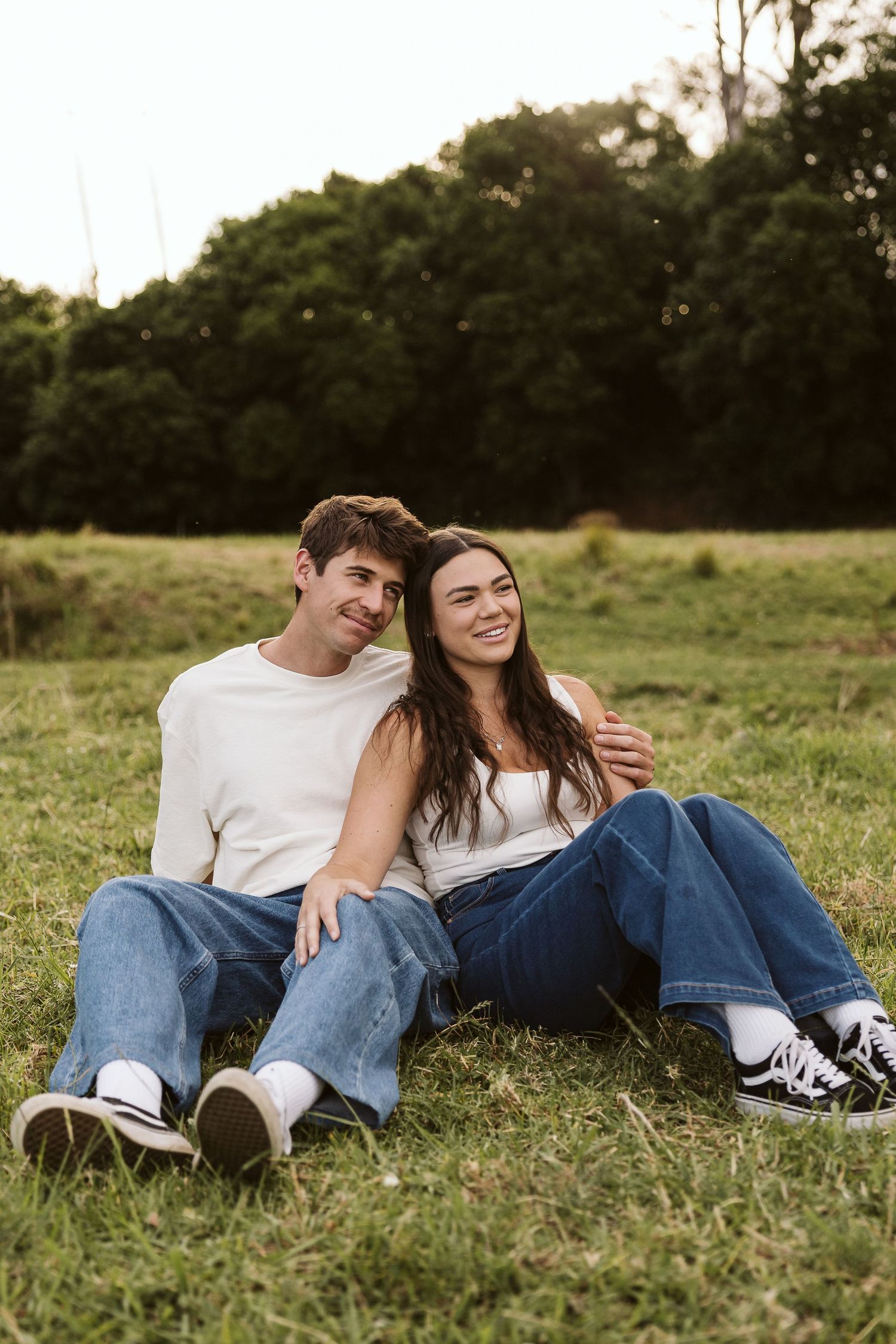 Happy couple sitting together on green grass field, both wearing white tops and blue jeans, smiling and embracing outdoors.