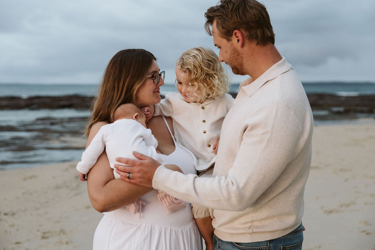 Young family on beach, mother holding newborn while toddler reaches toward father.