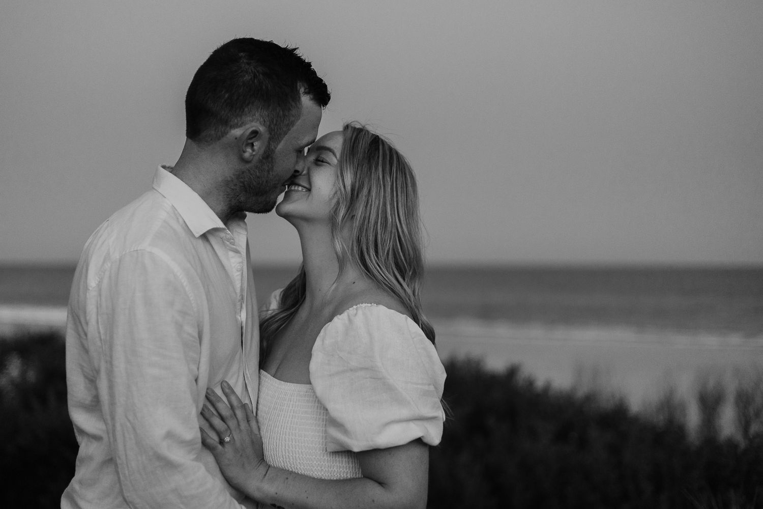 Black and white photo of couple smiling at each other with ocean in background.
