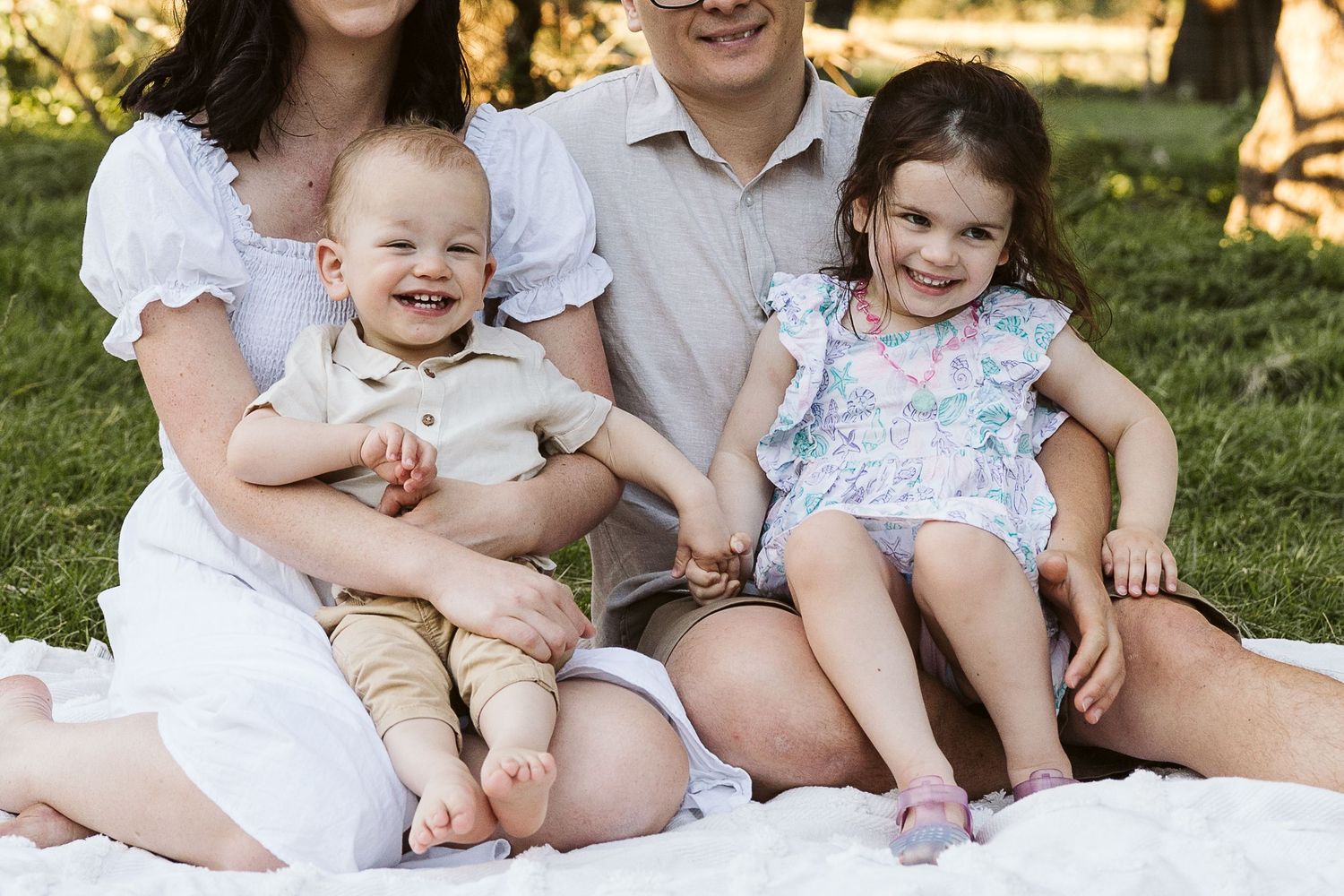 Happy family of four sitting on white blanket outdoors, parents with smiling baby boy and toddler girl in summer clothes.