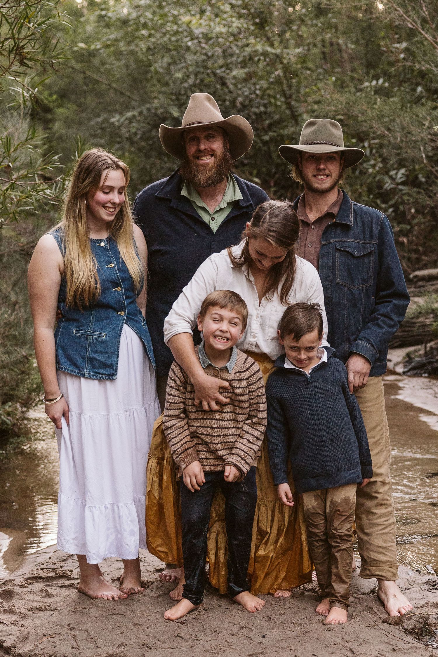 Family of six posing together barefoot by a creek in a natural bush setting.
