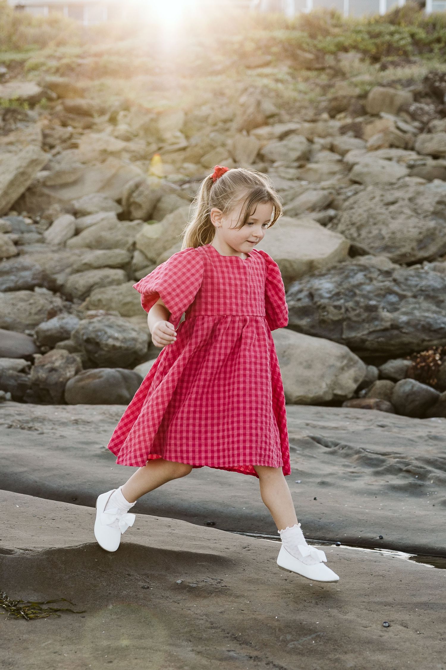 A young girl in a red gingham dress and white sneakers runs playfully on rocks with sunlight glowing behind her.