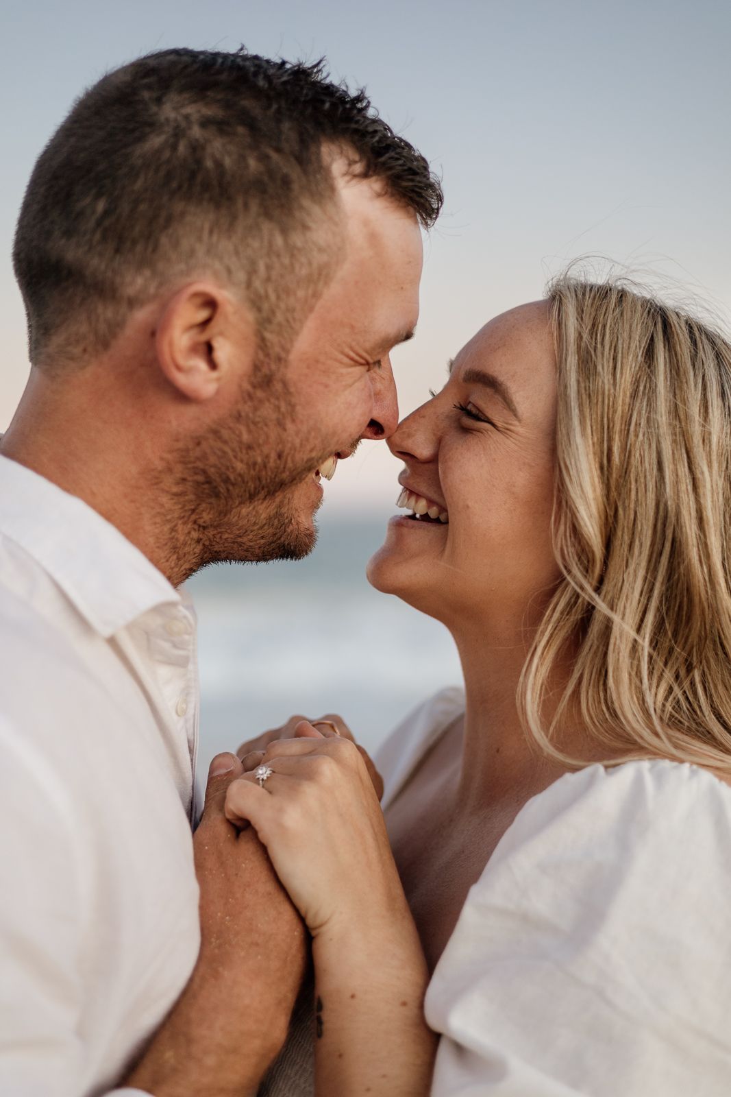Happy engaged couple smiling and holding hands at the beach, woman showing diamond engagement ring.