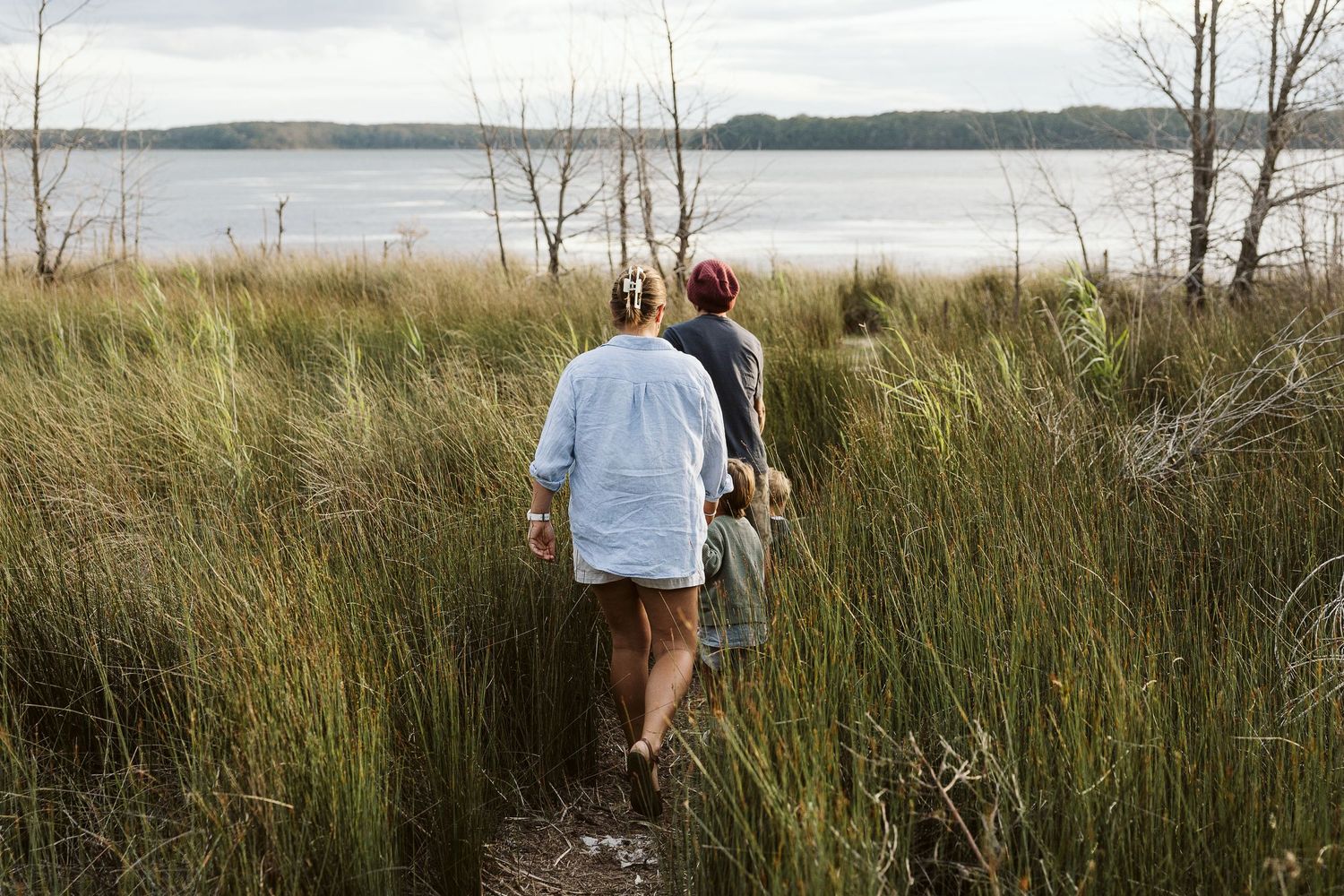 Family walking through tall grass toward a serene lake on a cloudy day.