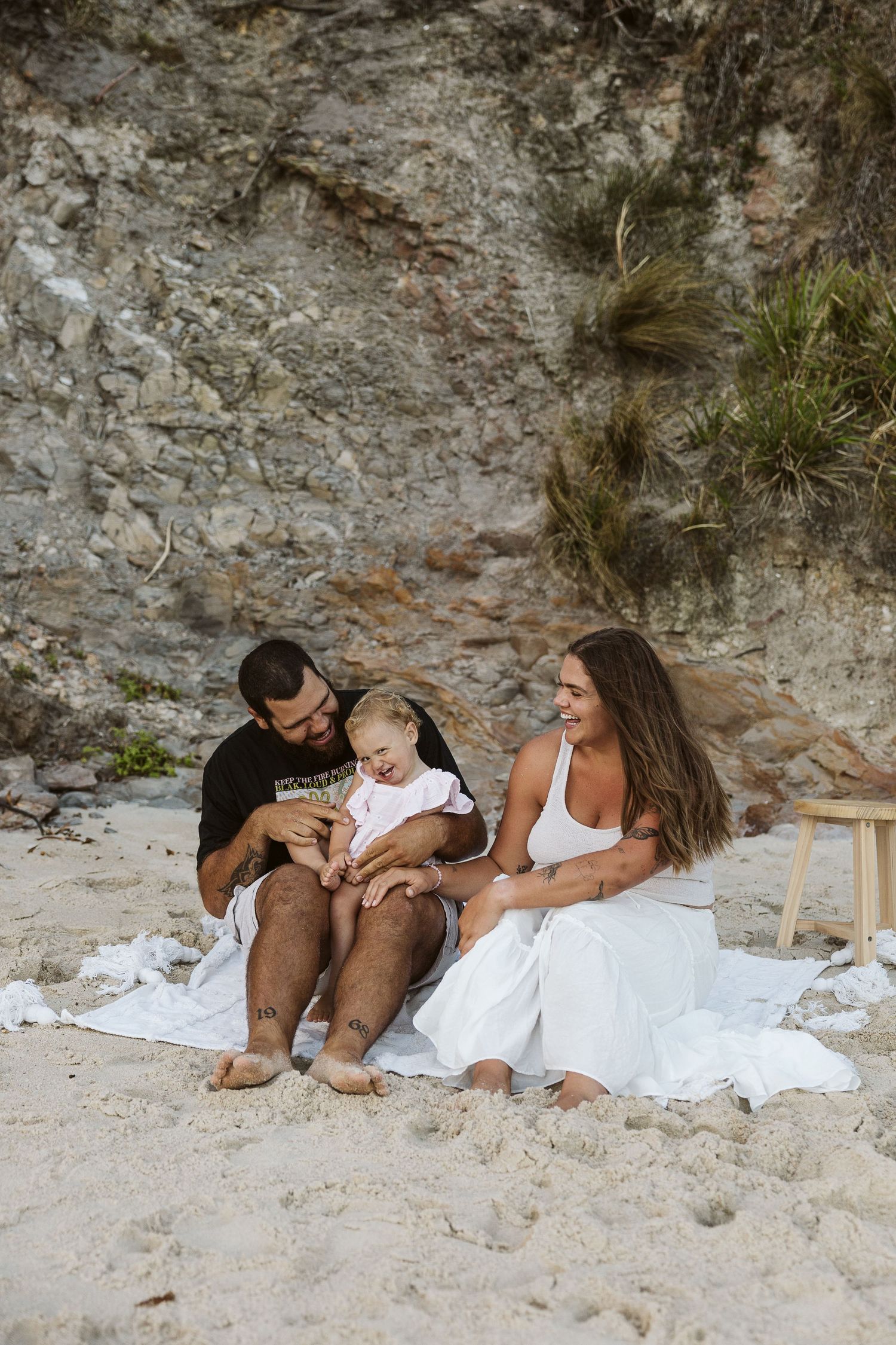 Family sitting on sandy beach with rocky cliff backdrop, parents smiling at baby in white onesie.
