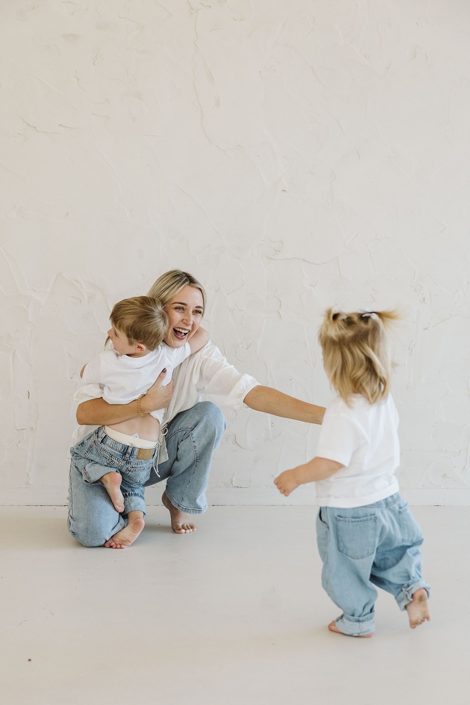 A laughing mother crouches to embrace her young son as her toddler daughter runs toward them, all wearing white shirts and jeans.