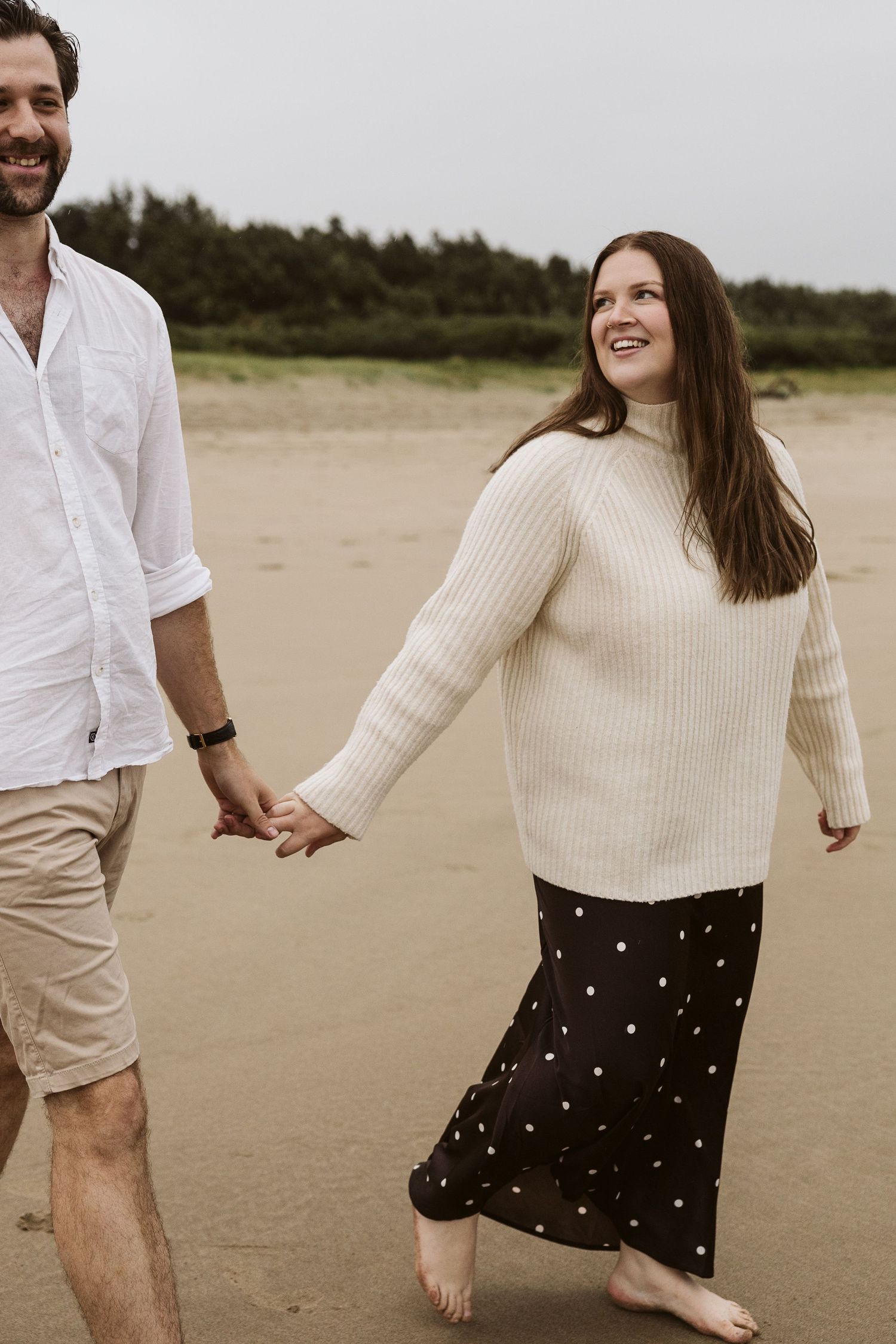 Couple holding hands and walking barefoot on a sandy beach, woman in cream sweater and polka dot skirt, man in white shirt.