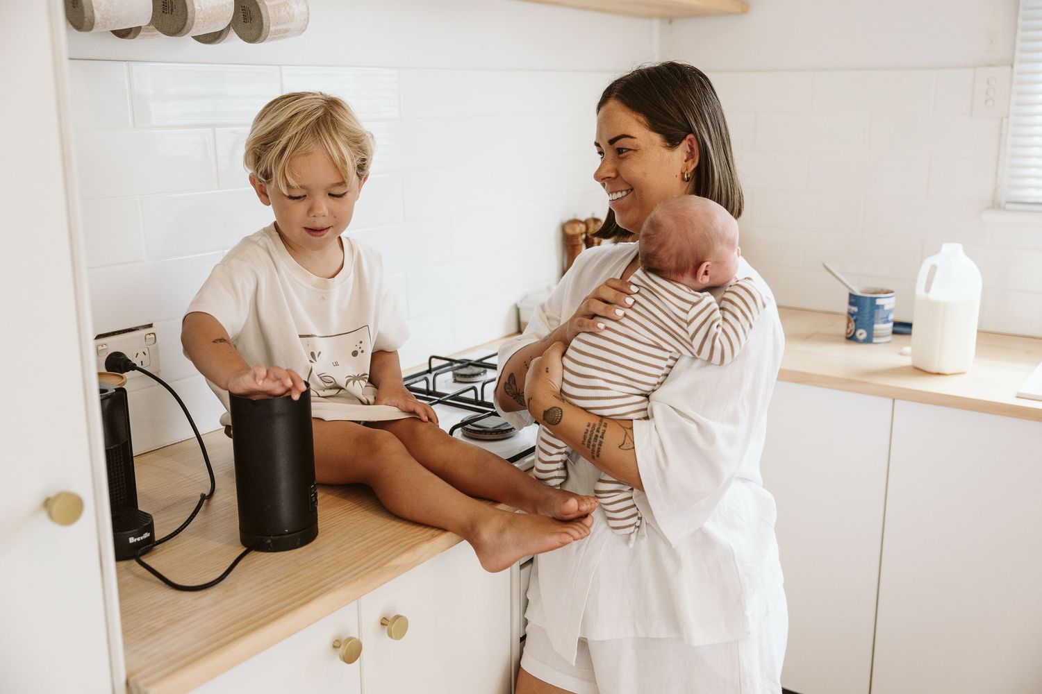 Mother in white robe holding newborn while toddler son sits on kitchen counter.