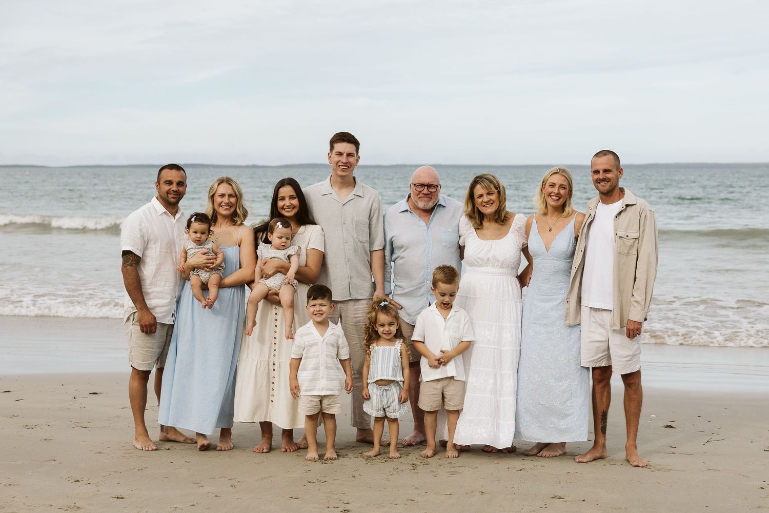 Large family group portrait on a sandy beach wearing coordinated white and blue outfits.