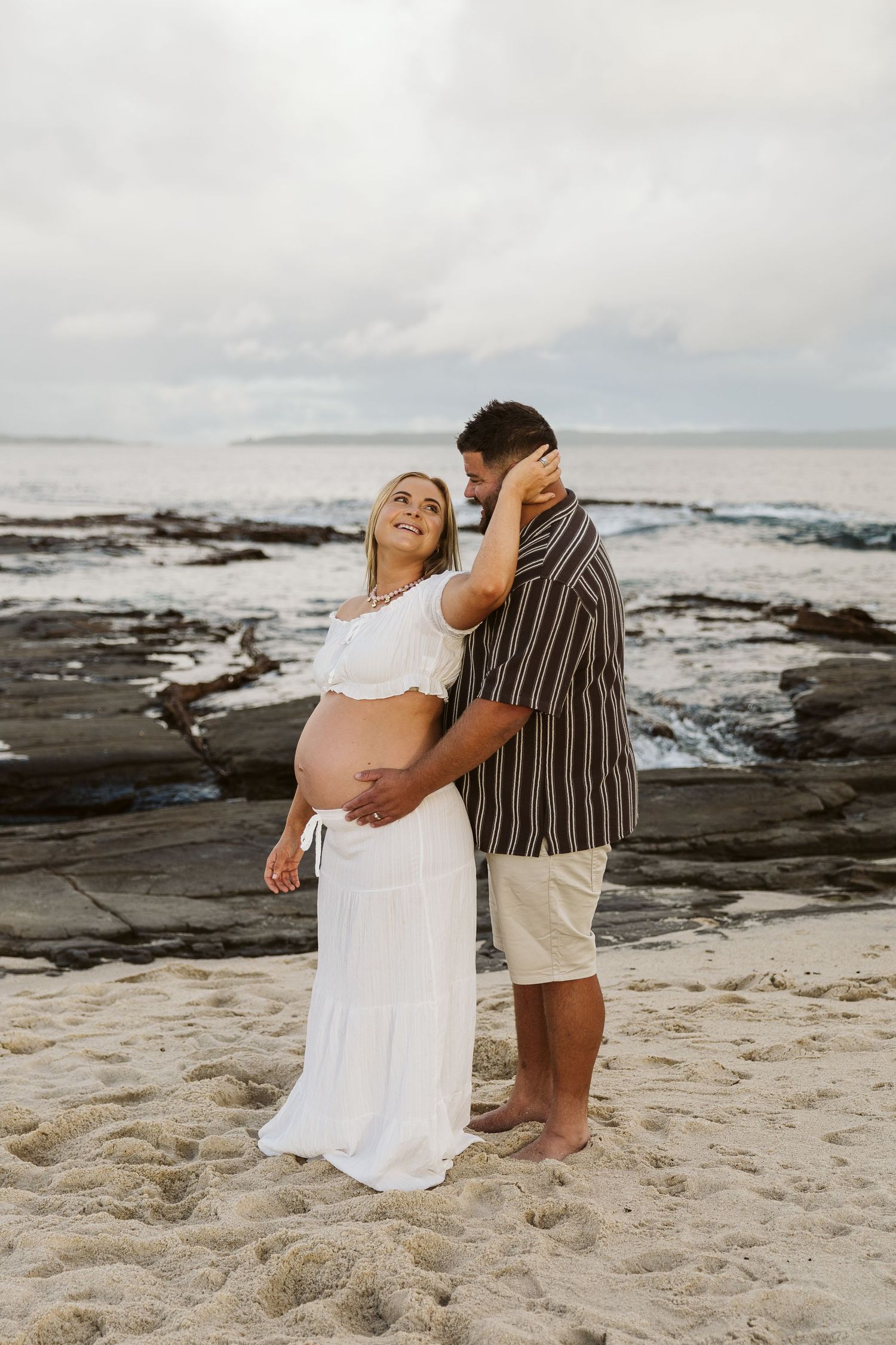 Pregnant woman in white outfit embraces partner on sandy beach by ocean.