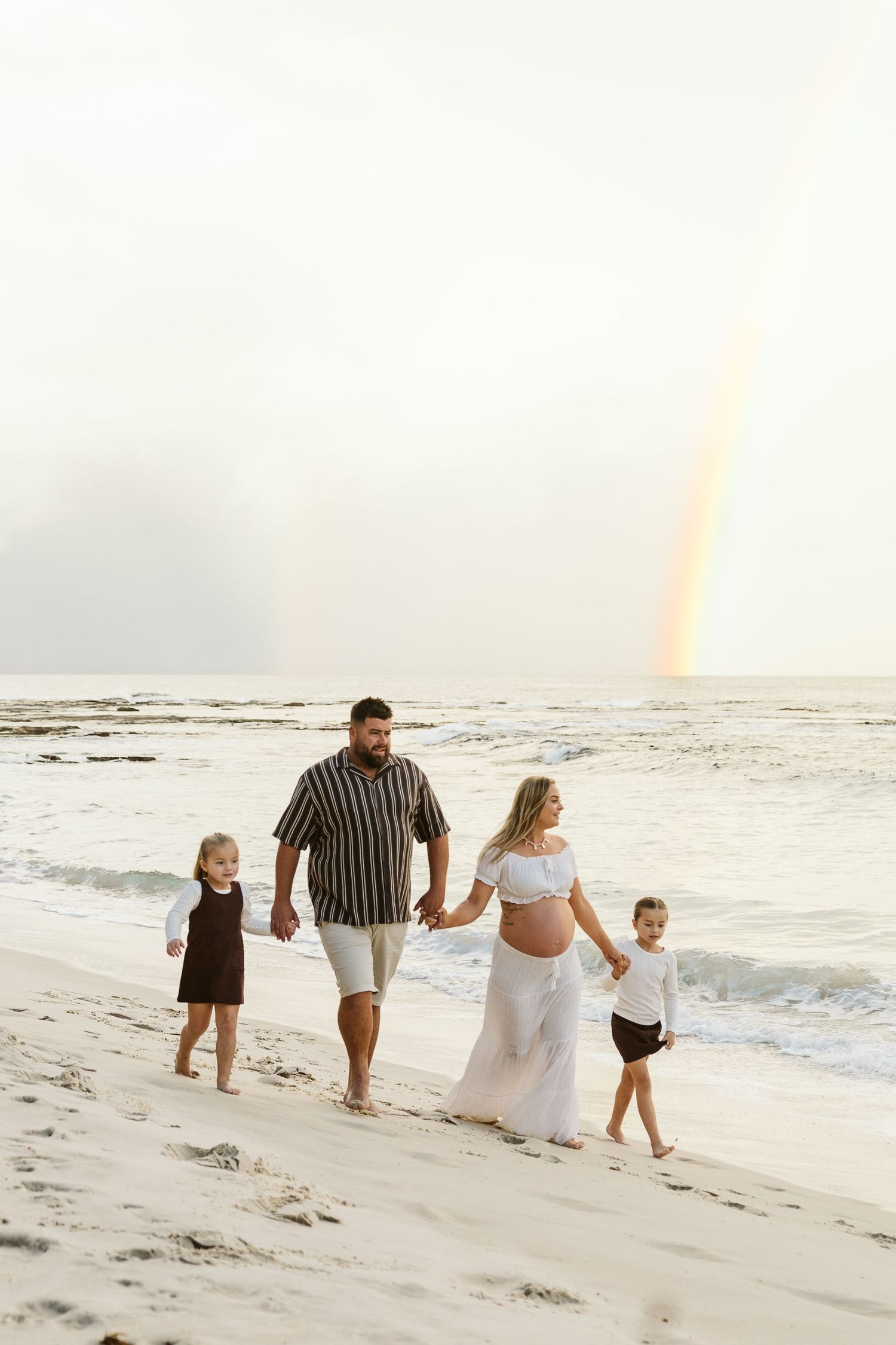 Pregnant woman and family holding hands walking along a white sandy beach with a rainbow in the sky.