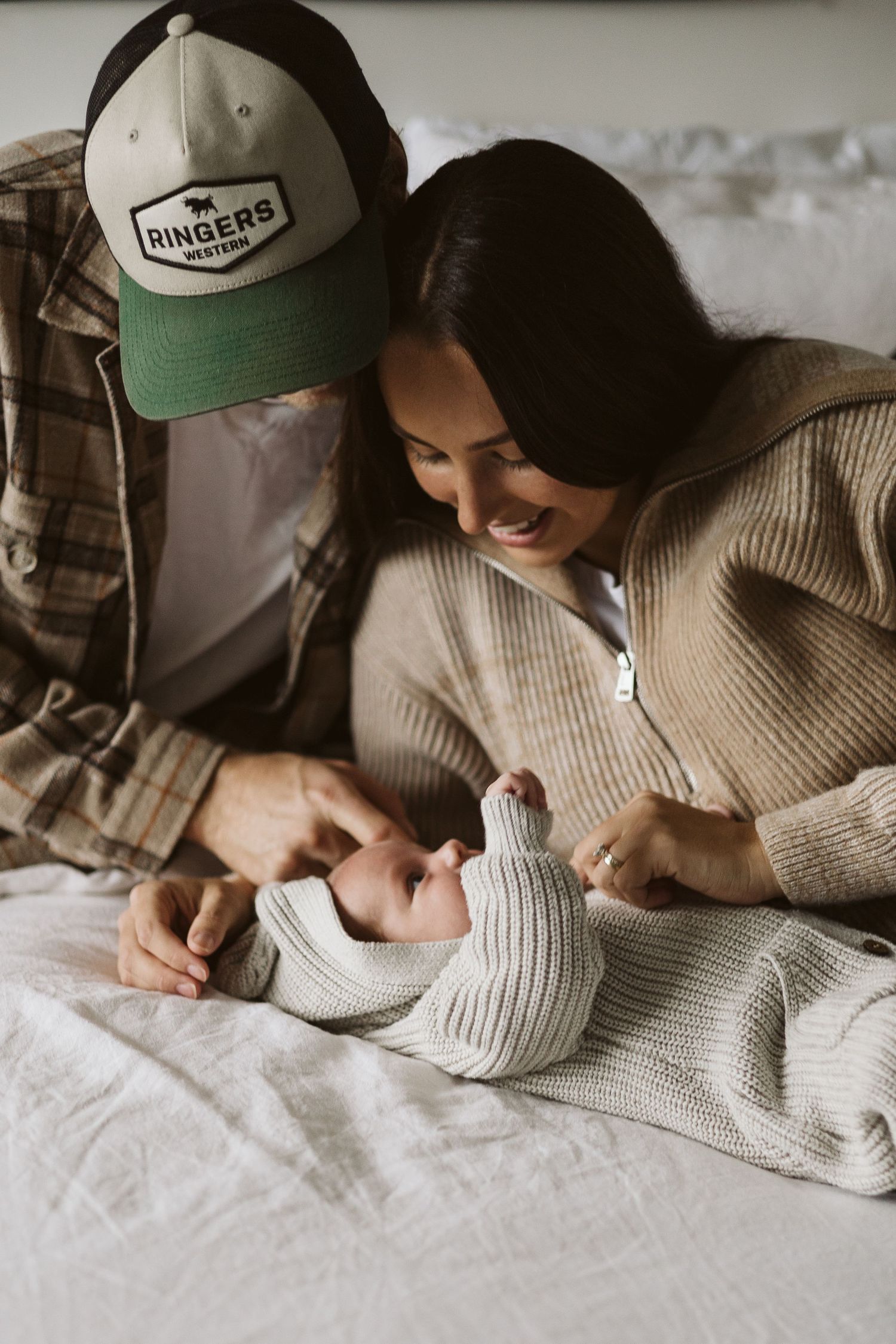 Happy parents in casual wear smile down at their newborn lying on a bed.