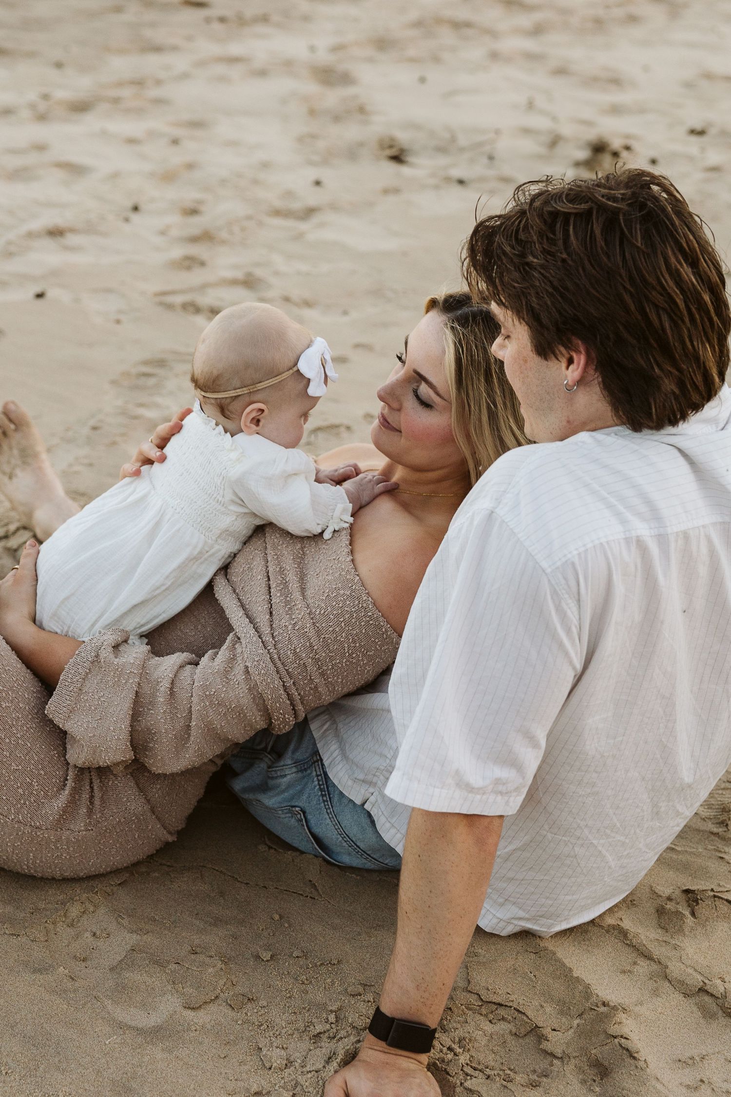 Young family sitting on sandy beach, mother holding baby in white dress while father looks on lovingly.